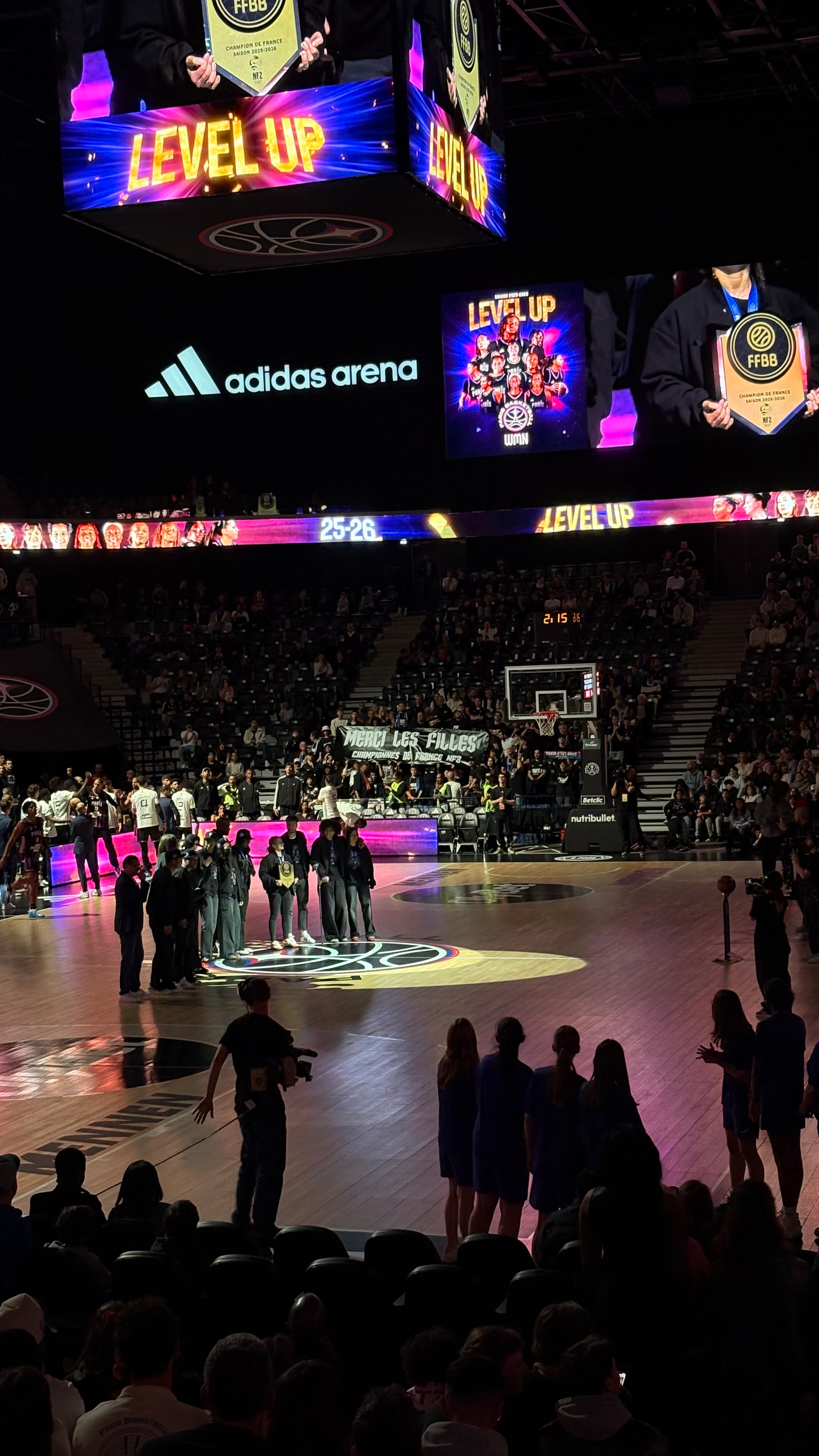 A basketball court in an arena is the setting for a ceremony involving a group of people, with LEVEL UP displayed on screens above.
