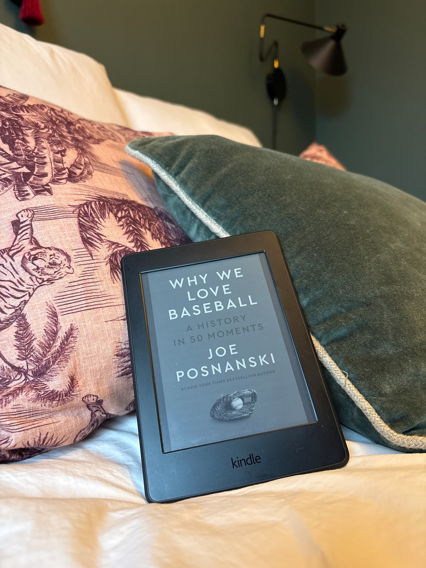A Kindle displaying the book Why We Love Baseball by Joe Posnanski rests on a bed with patterned pillows and a lamp in the background.