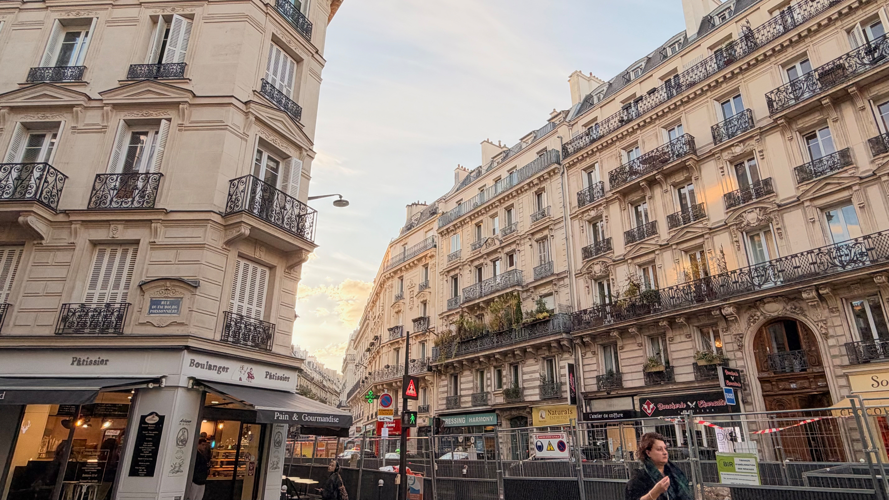 A person is walking on a street lined with classic Parisian buildings featuring balconies and shops at street level during the early evening.