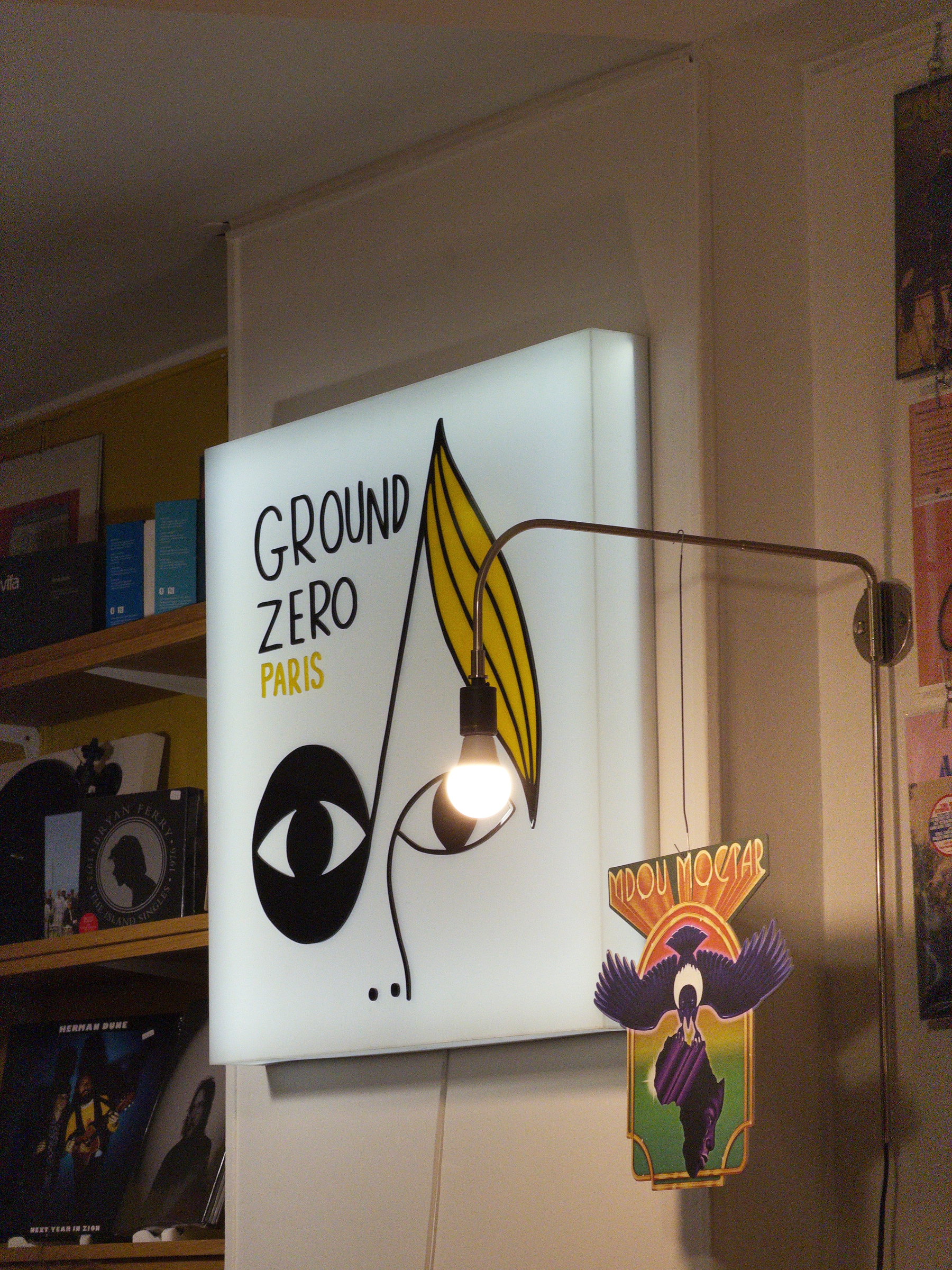 A glowing sign with the text GROUND ZERO PARIS and an abstract face drawing is mounted on a wall next to a hanging light bulb and a decorative card.