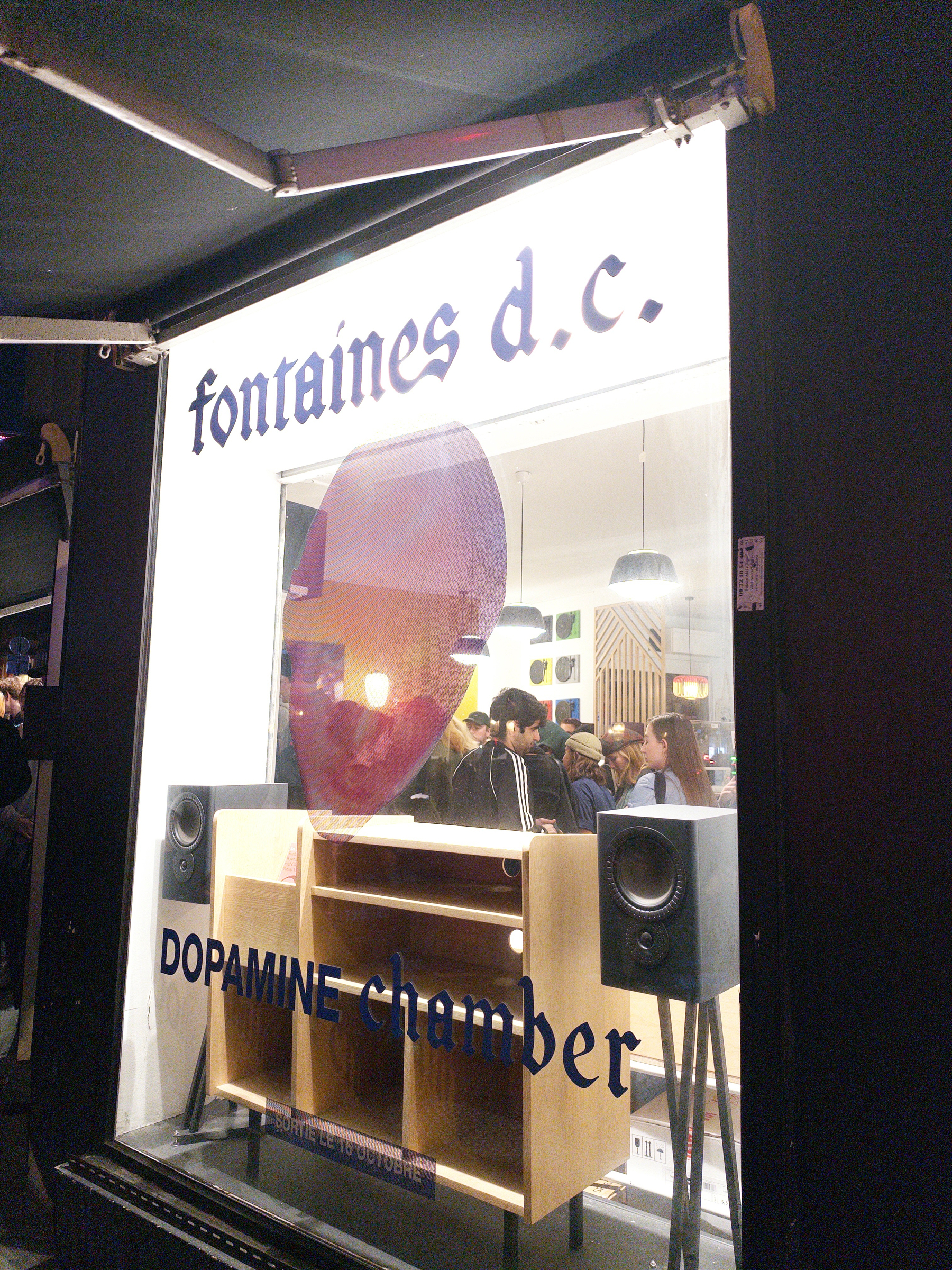 A group of people is gathered inside a brightly lit space with the words fontaines d.c. and DOPAMINE chamber displayed on the window.