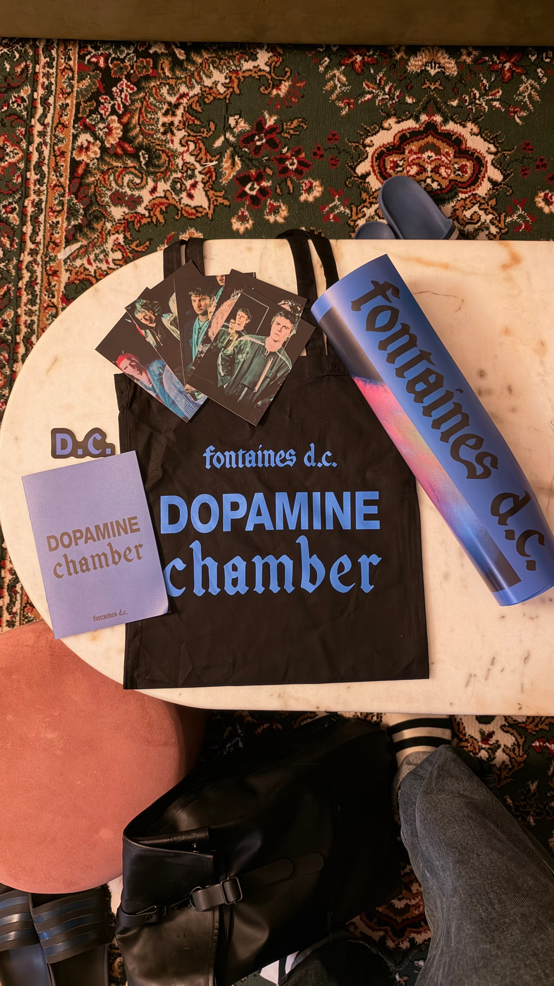 A collection of Fontaines D.C. merchandise including a tote bag, postcards, a booklet titled Dopamine Chamber, and a rolled-up poster is displayed on a marble table.