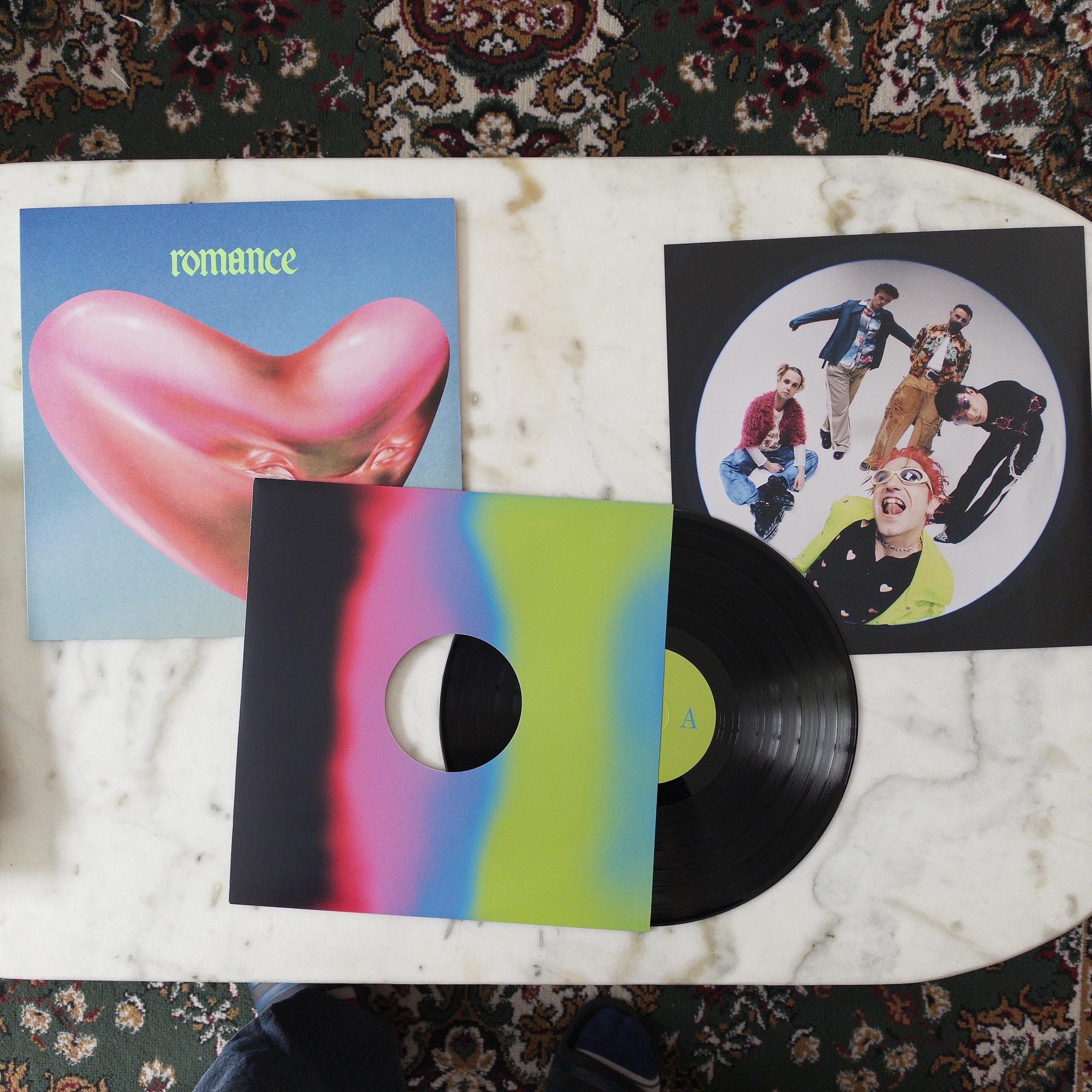 A vinyl record (Romance by the band Fontaines D.C.) and its colorful sleeves, including one with the word romance and a heart-shaped pink object, are displayed on a marble surface.