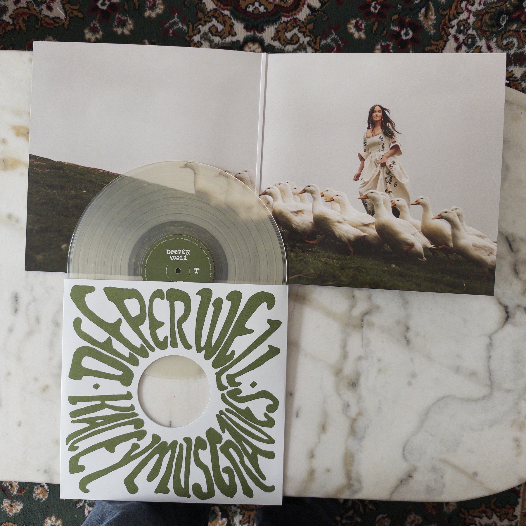 A clear vinyl record titled Deeper Well by Kacey Musgraves is partially removed from its sleeve, which features a photograph of Haley Musgraves standing on a grassy hill surrounded by white geese.