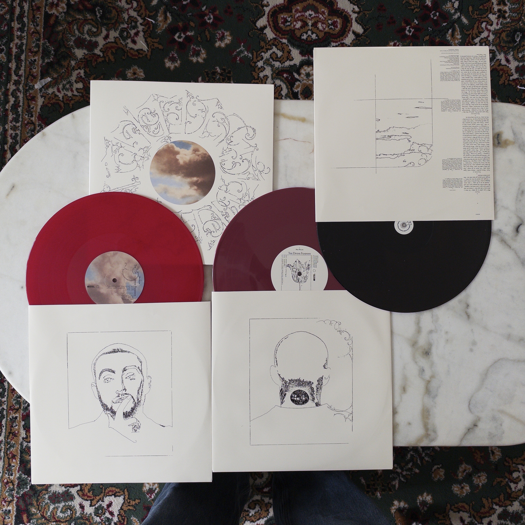 Three vinyl records (The Divine Feminine 10th Anniversary, by Mac Miller), two maroon and two black, are arranged on a white marble table, each with detailed line art on their sleeves, with the central sleeve depicting a head with intricate designs and a cloudy sky artwork.