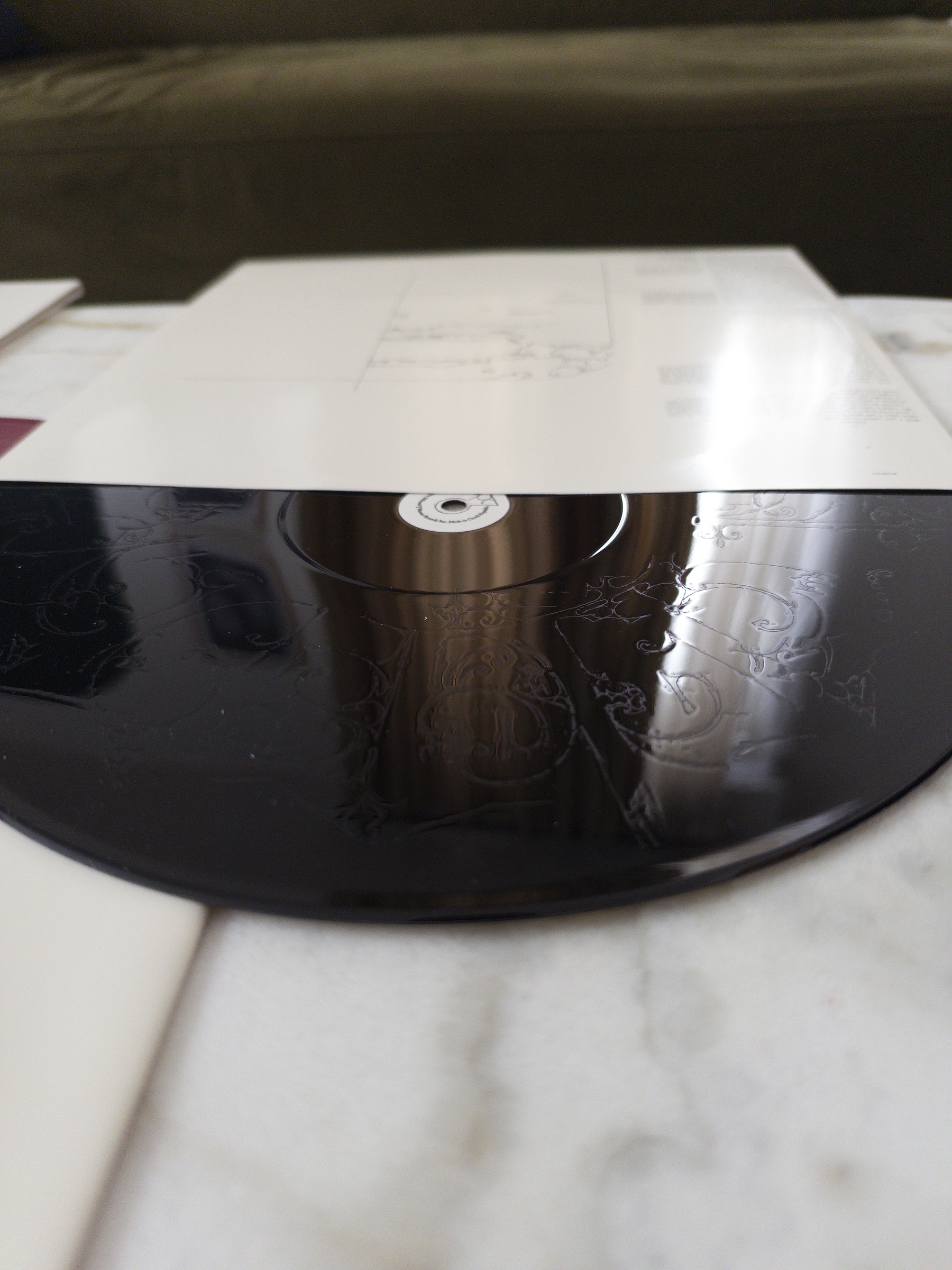 A vinyl record with subtle, intricate etched designs is placed on a white surface with papers or album covers in the background.