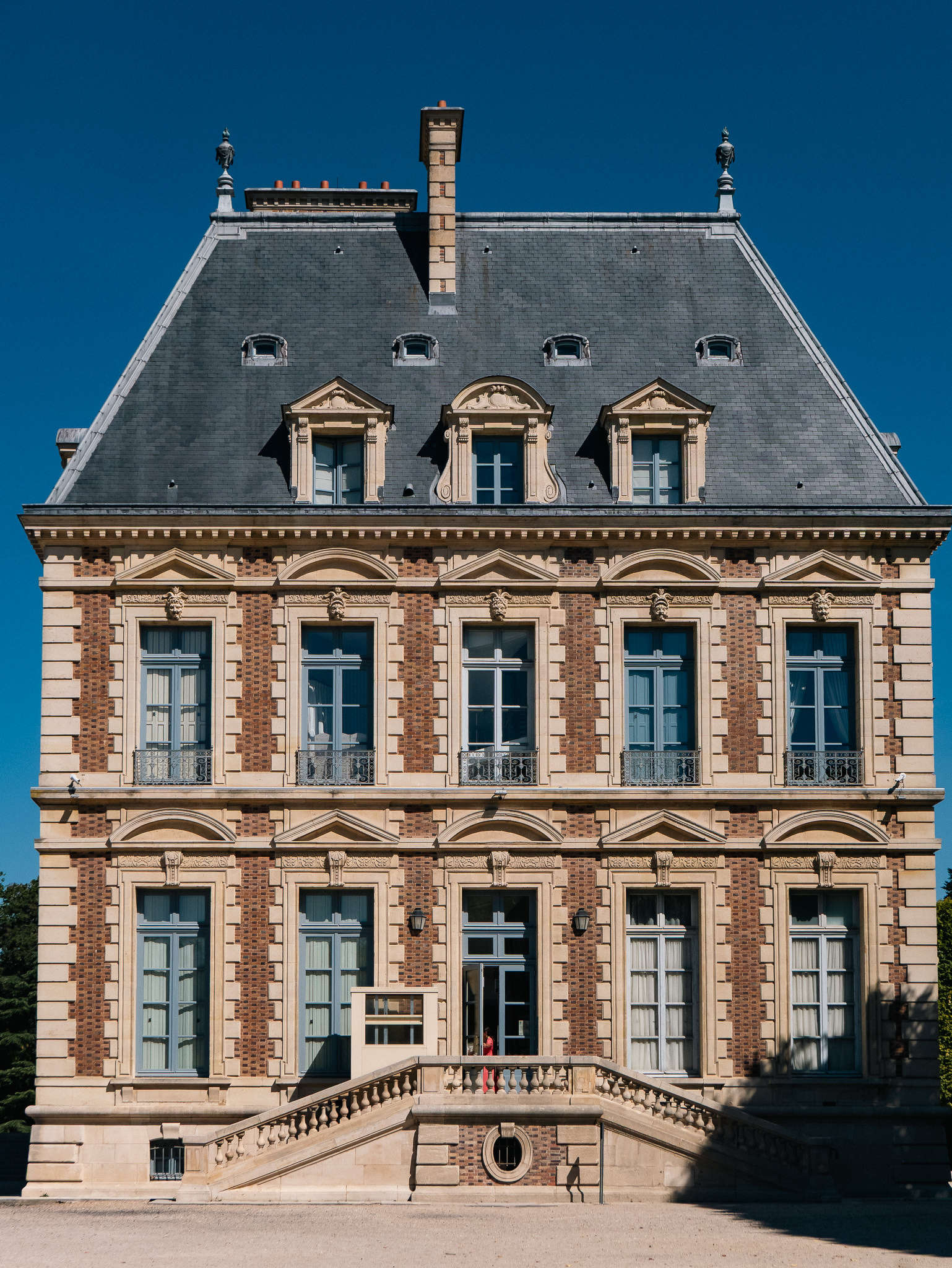 Auto-generated description: A grand, historic building with a slate roof, tall windows, ornate brickwork, and a central staircase leading to an entrance is set against a clear blue sky.