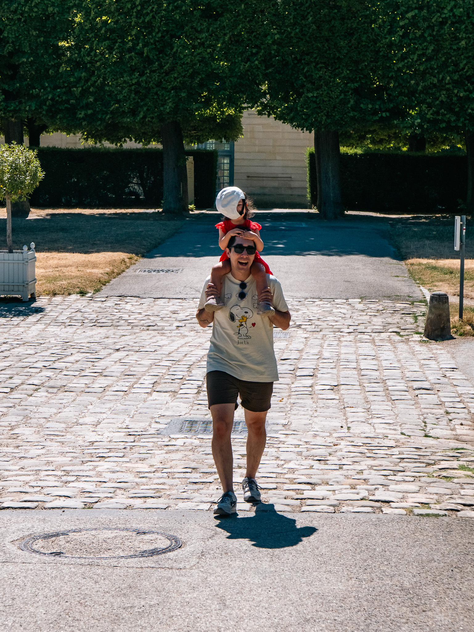 Auto-generated description: A man wearing sunglasses carries a child dressed in red on his shoulders while walking on a cobblestone path with trees and greenery in the background.
