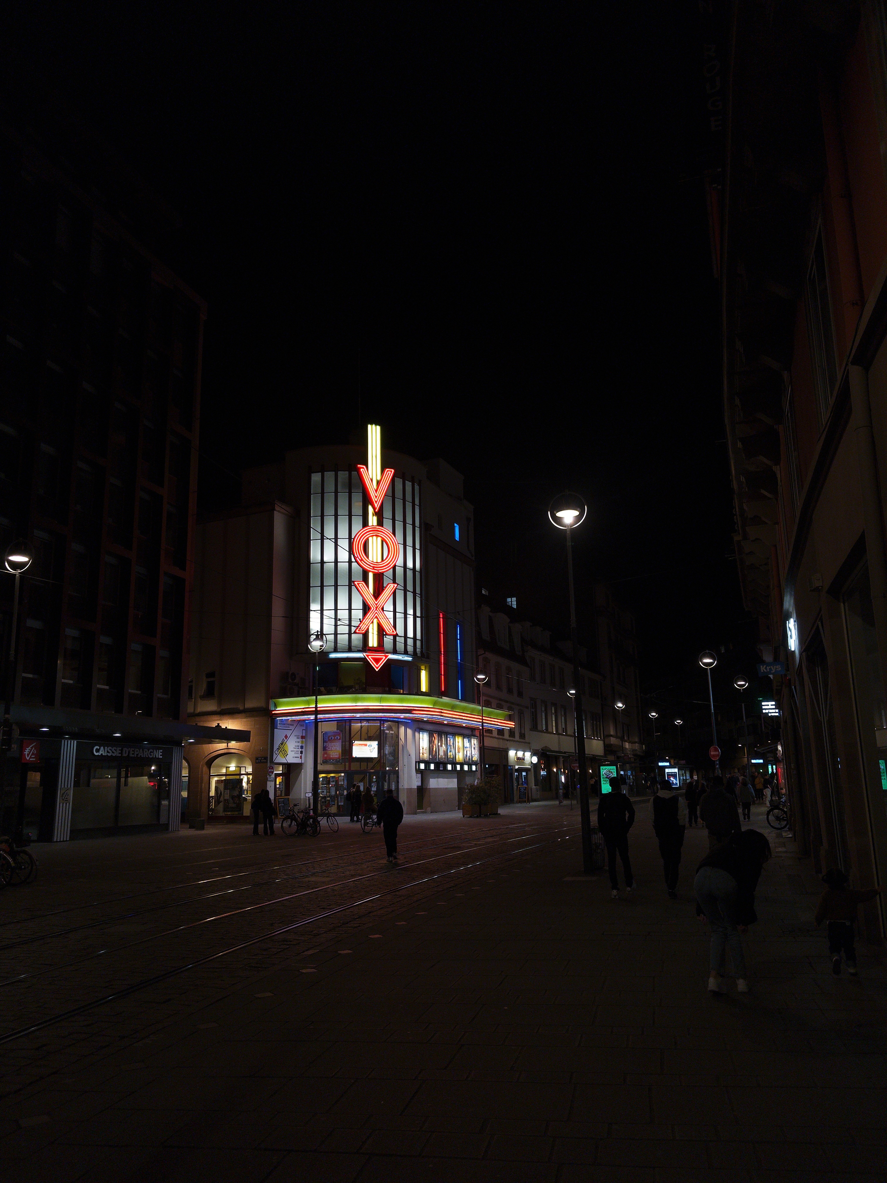 A street scene at night showcases a brightly lit VOX sign atop a building, with people walking along the sidewalk.