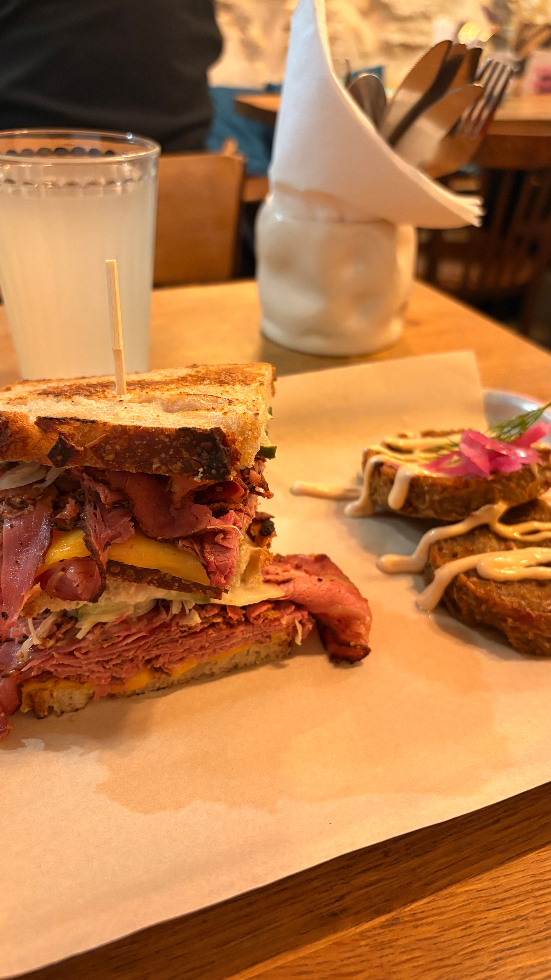 Auto-generated description: A sandwich stacked with meat and a side of seasoned fries is served on a wooden table alongside a glass of lemonade.