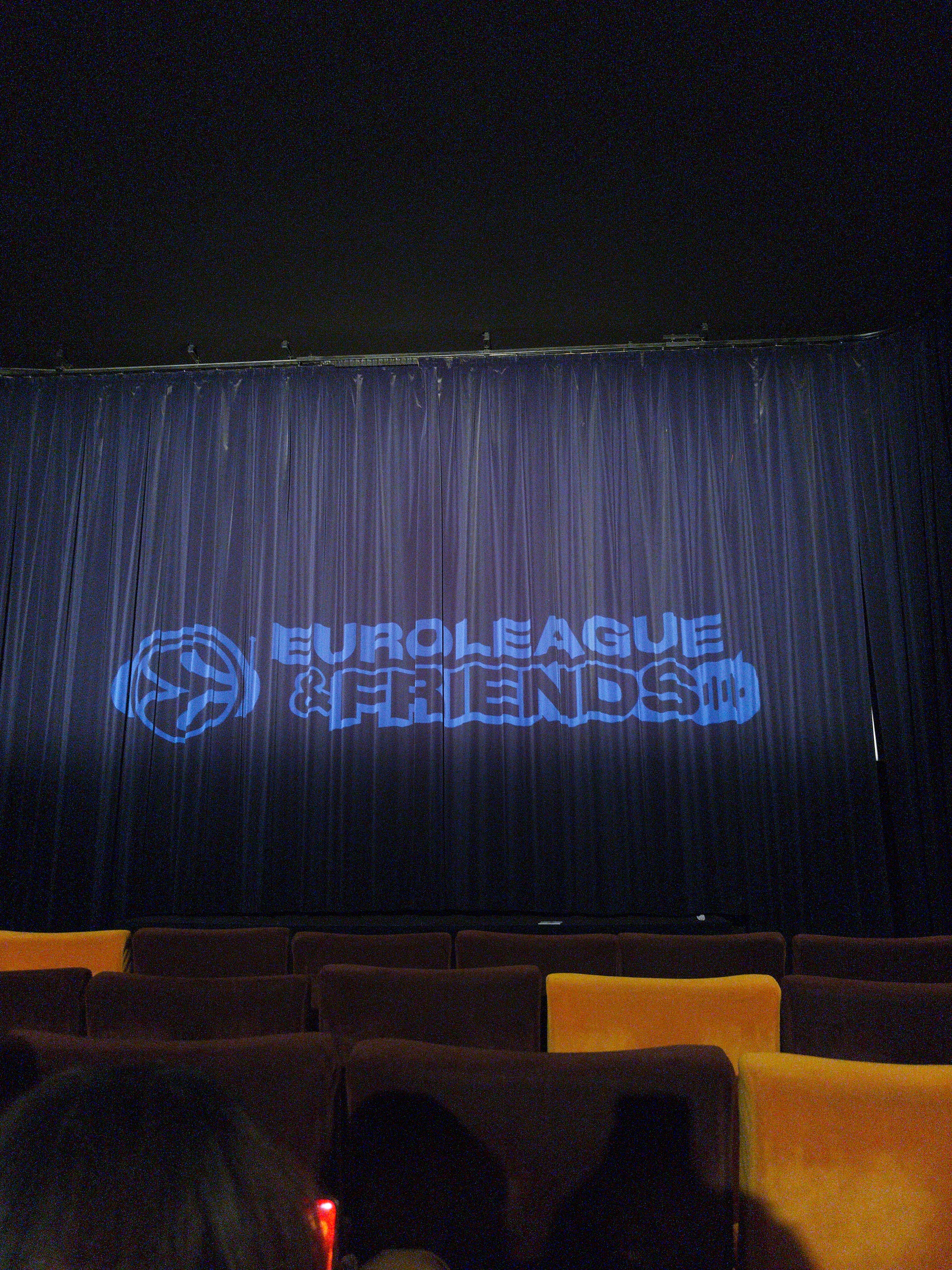 A dark theater room with a blue curtain displays the projection Euroleague & Friends above rows of empty brown seats.