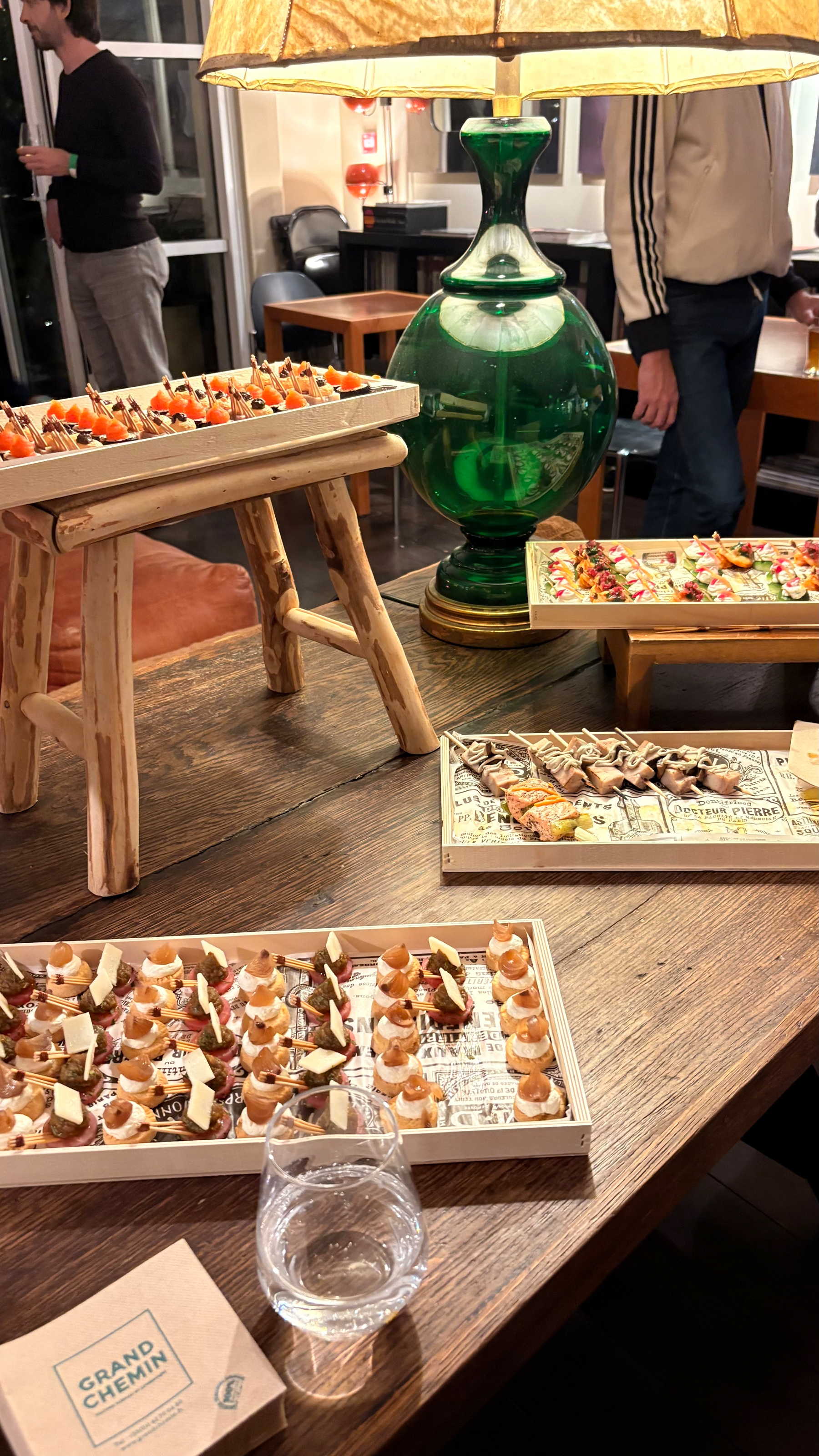 A wooden table displays assorted colorful appetizers and a green lamp, with people socializing in the background.