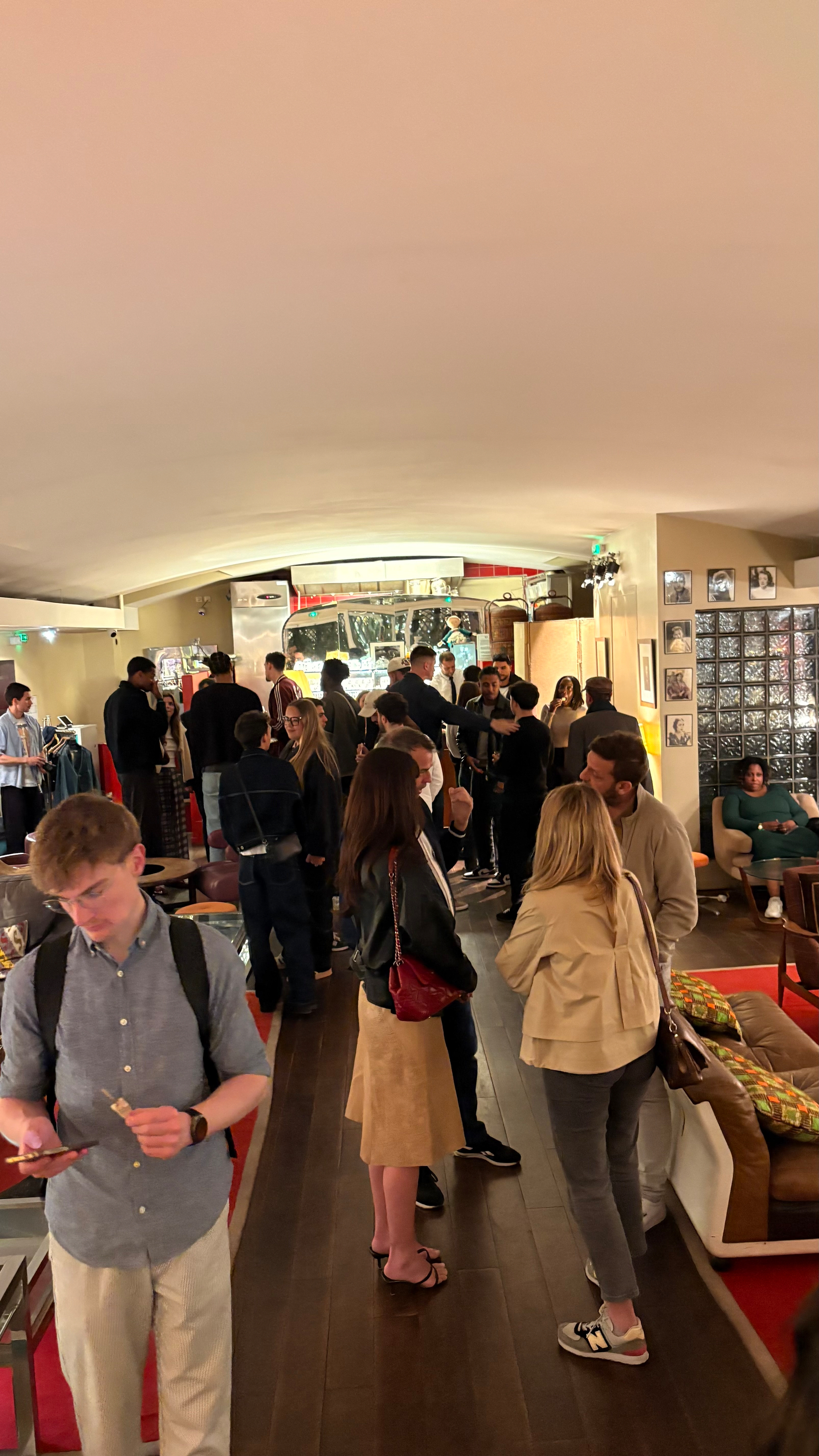 A group of people is socializing in a warmly lit room with couches and a bar area in the background.