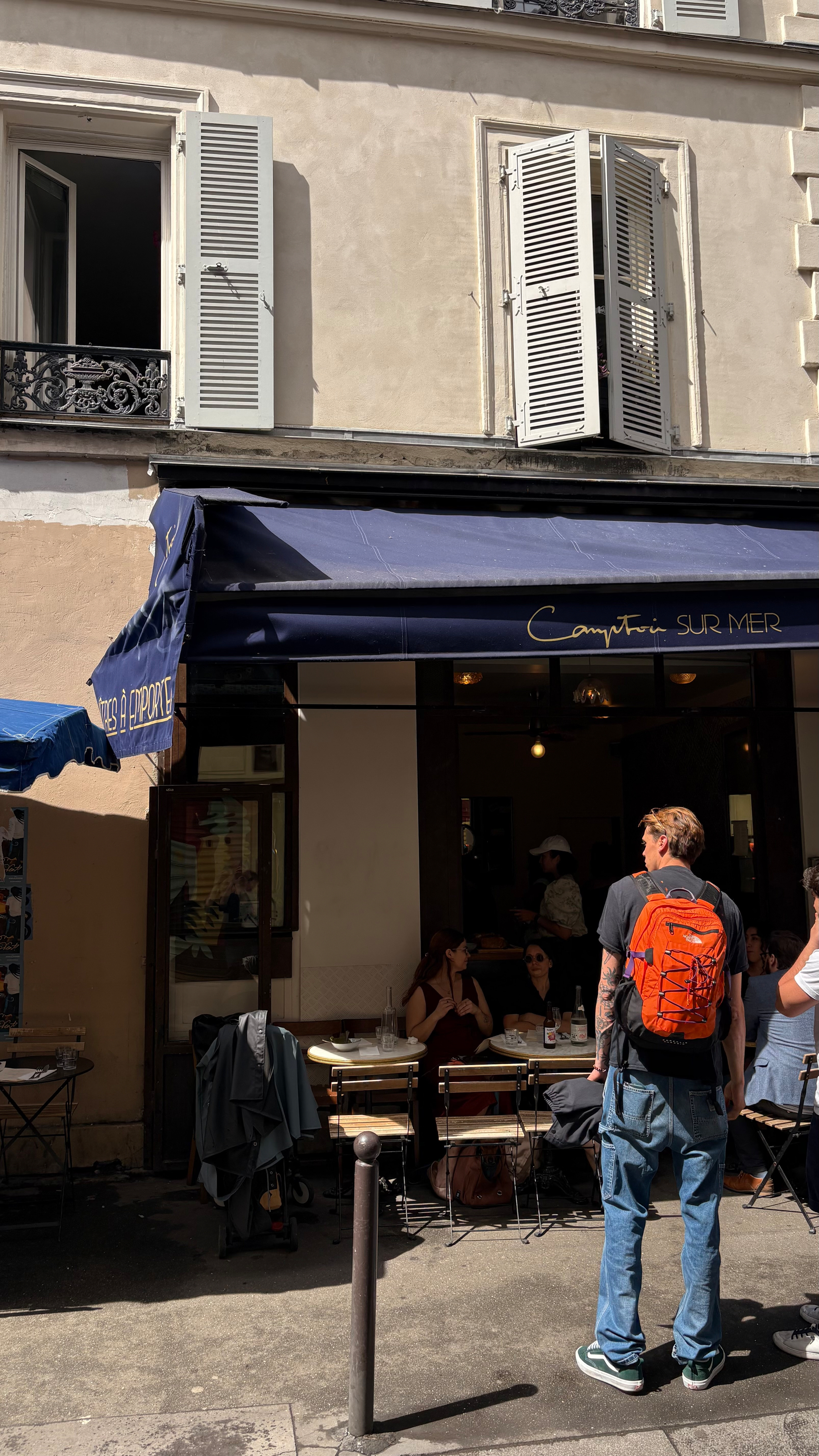 People are gathered outside a quaint café with a navy blue awning on a sunny day.