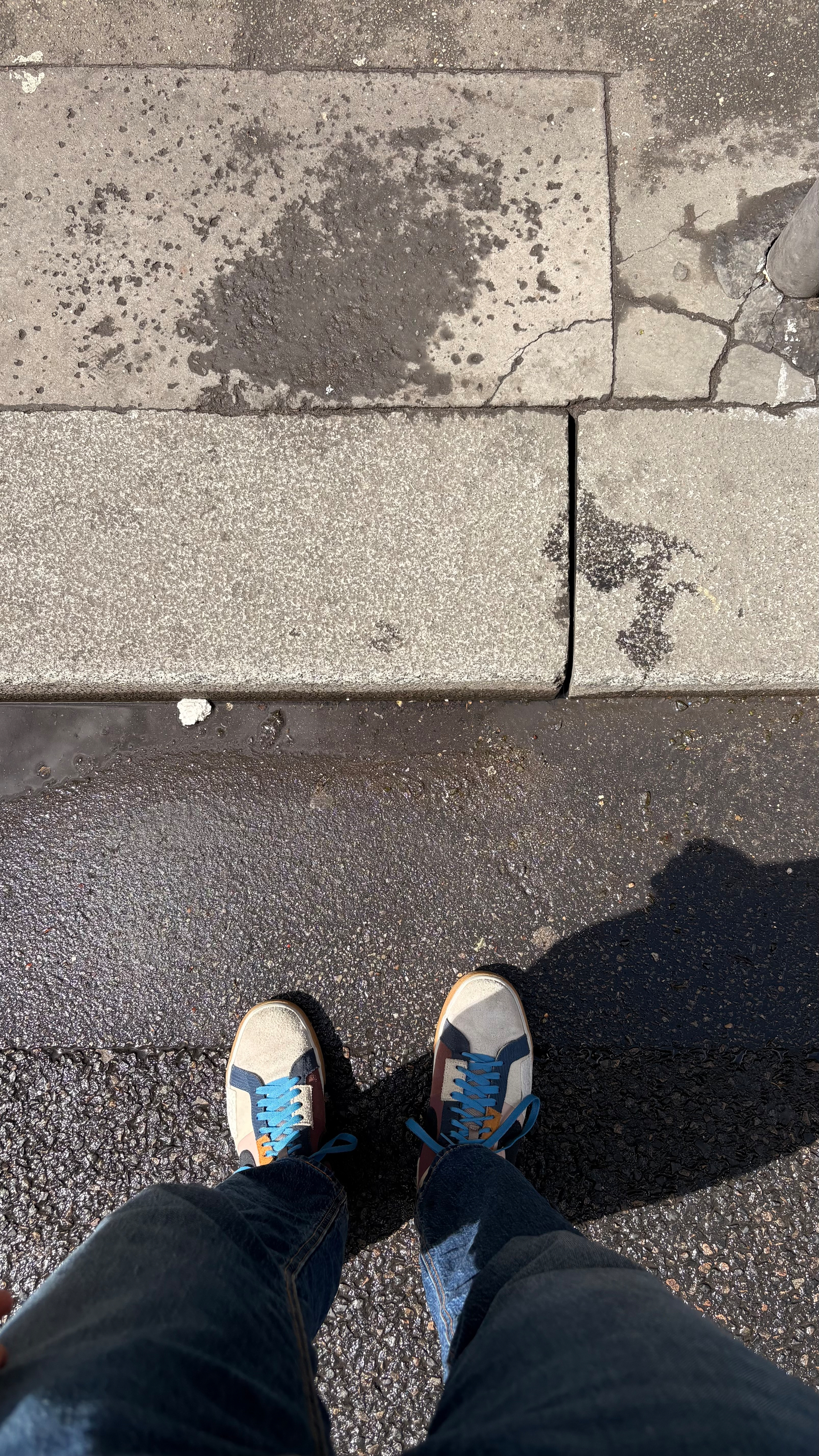 A person wearing blue jeans and sneakers with blue laces is standing on a textured sidewalk.
