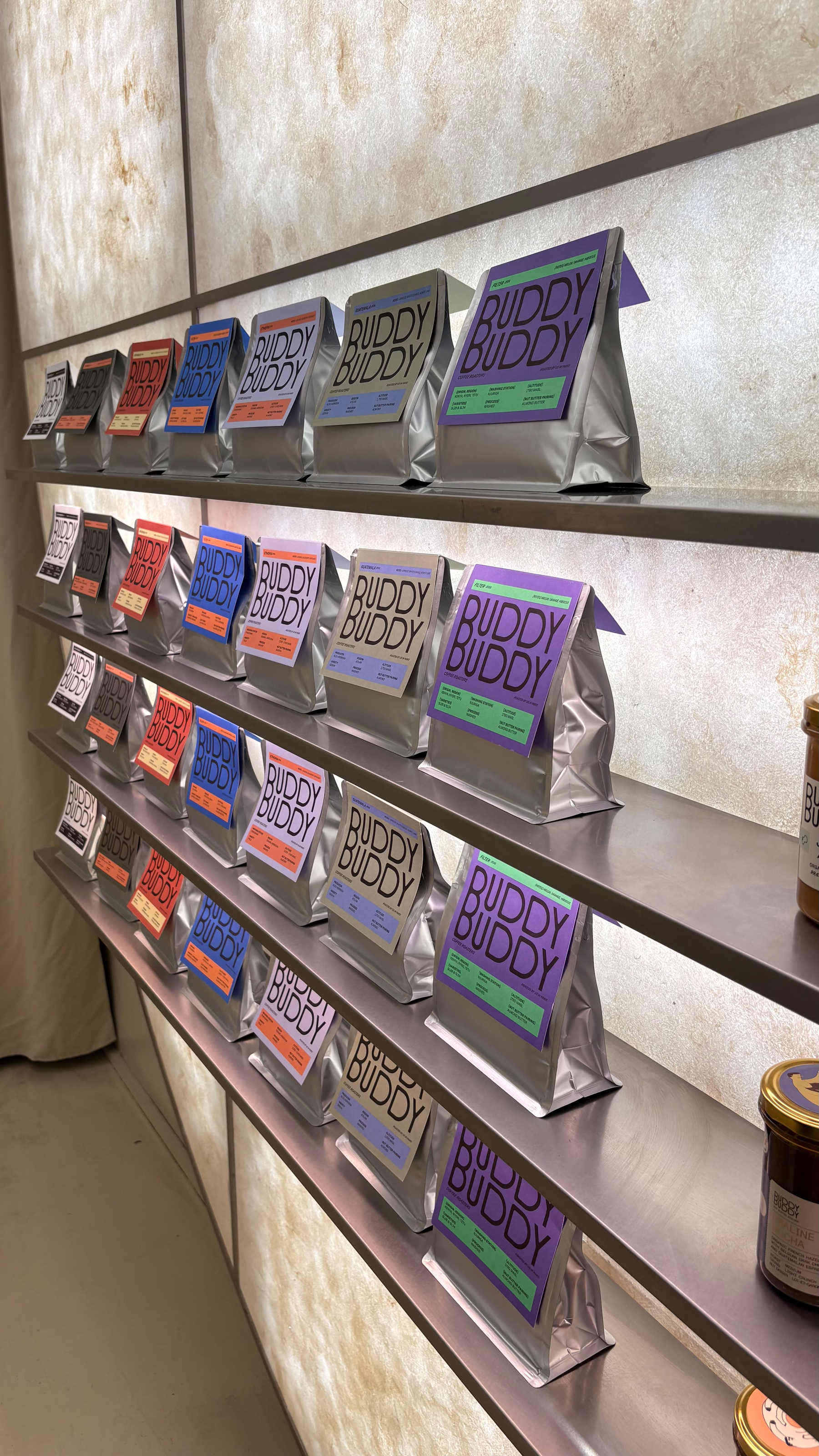 Packets of various flavors of BUDDY BUDDY products are neatly displayed on shelves against a softly lit background.
