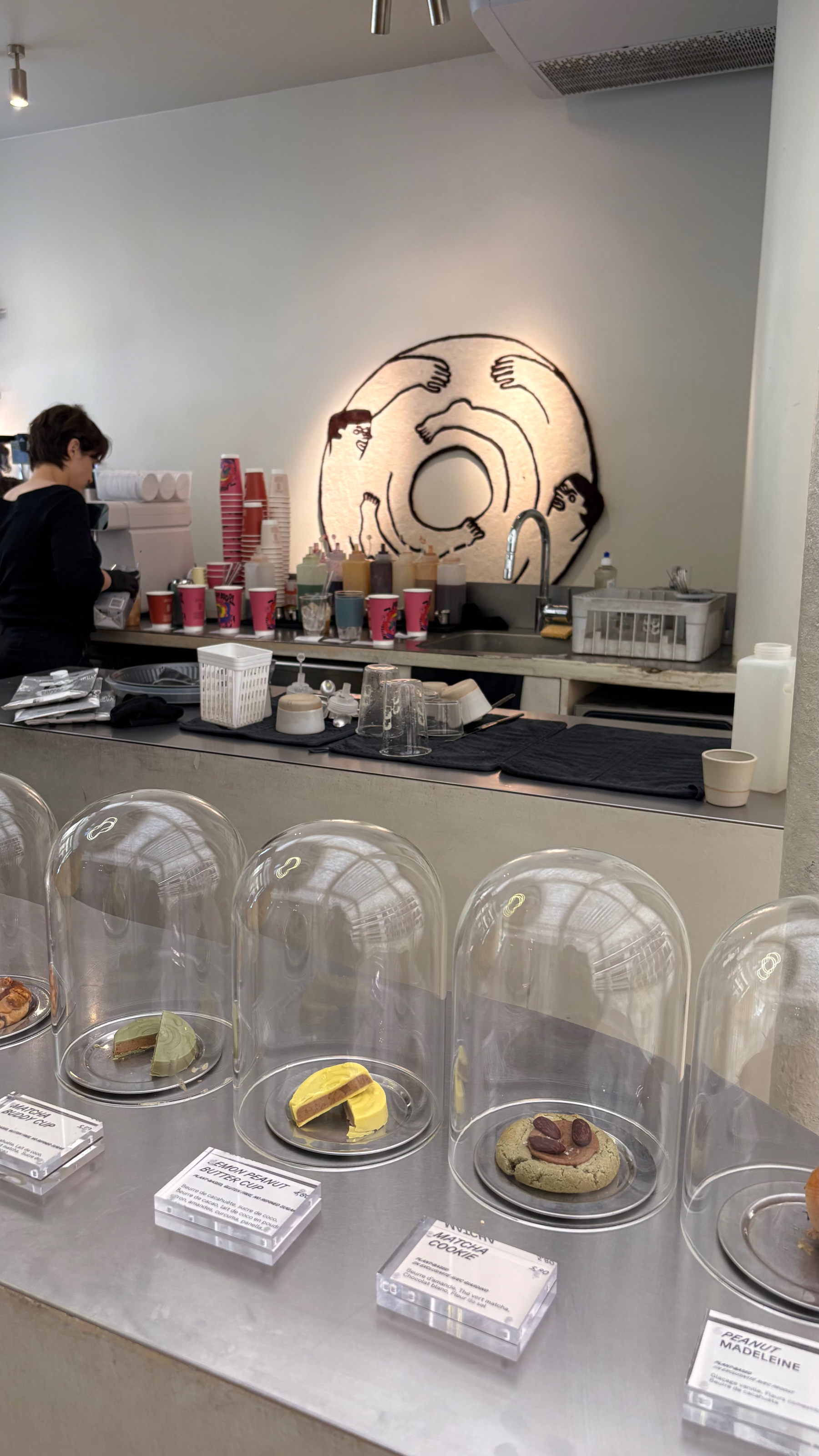 A cafe counter displays assorted pastries under glass domes, with a barista preparing drinks and a wall mural depicting people enjoying a large doughnut.