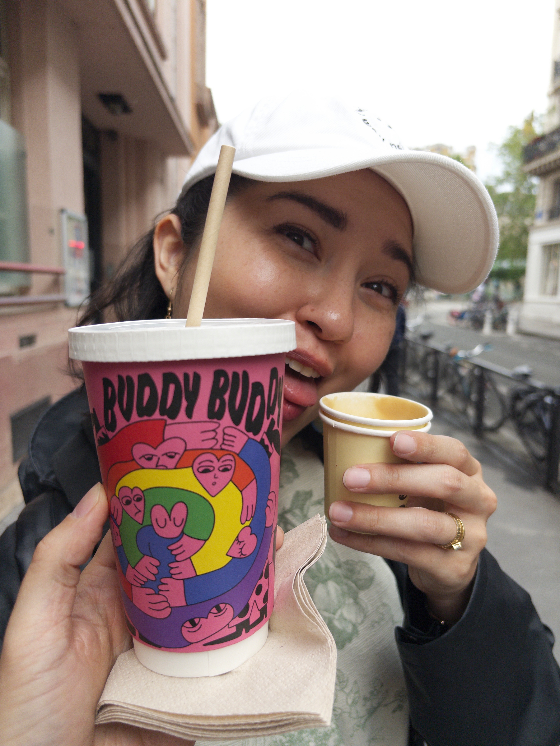 A person in a white cap holds a colorful cup labeled BUDDY BUDDY and another smaller cup, smiling on a city street.