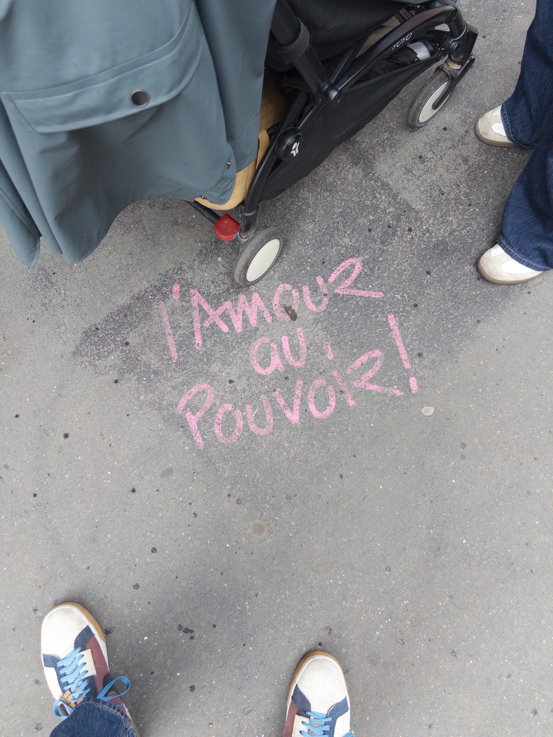 Pink chalk on the pavement spells l'Amour au Pouvoir! surrounded by people wearing shoes and a stroller.
