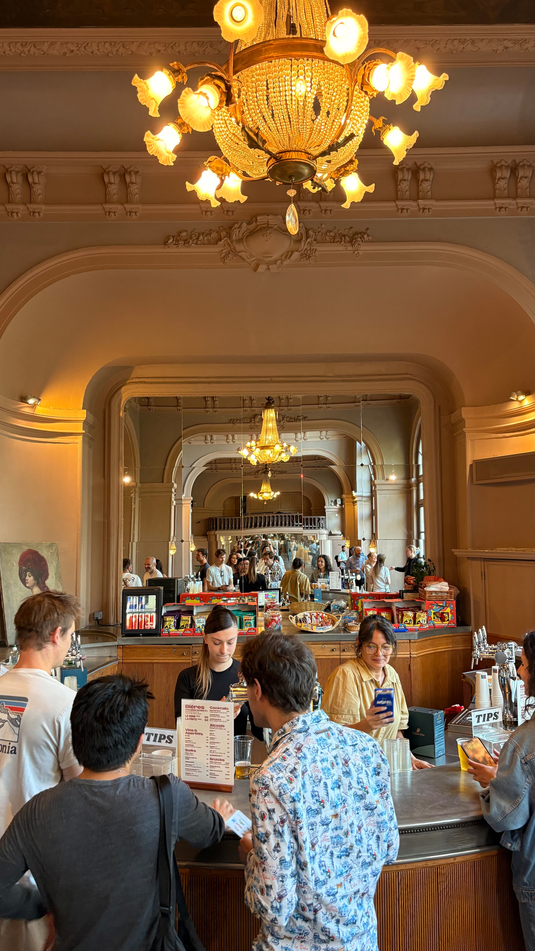 A group of people are gathered around a bar counter in a grand, ornately decorated room with chandeliers and mirrors.