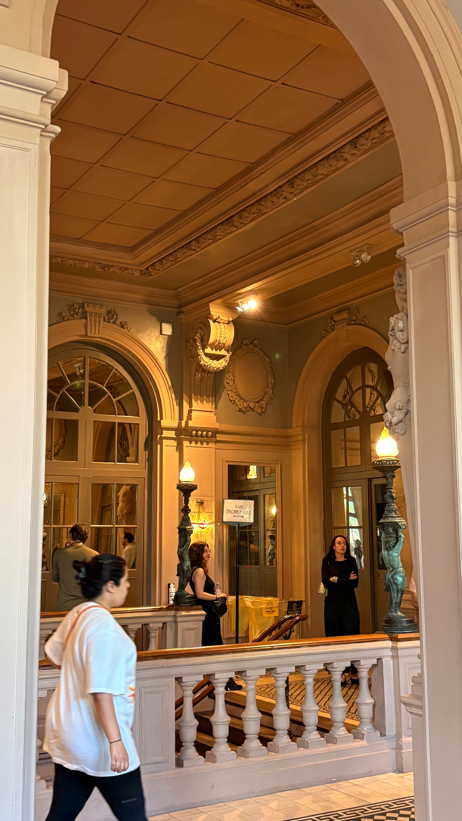 An elegant indoor setting features arched doorways, intricate wall designs, ornate lamps, and individuals standing and walking through the space.