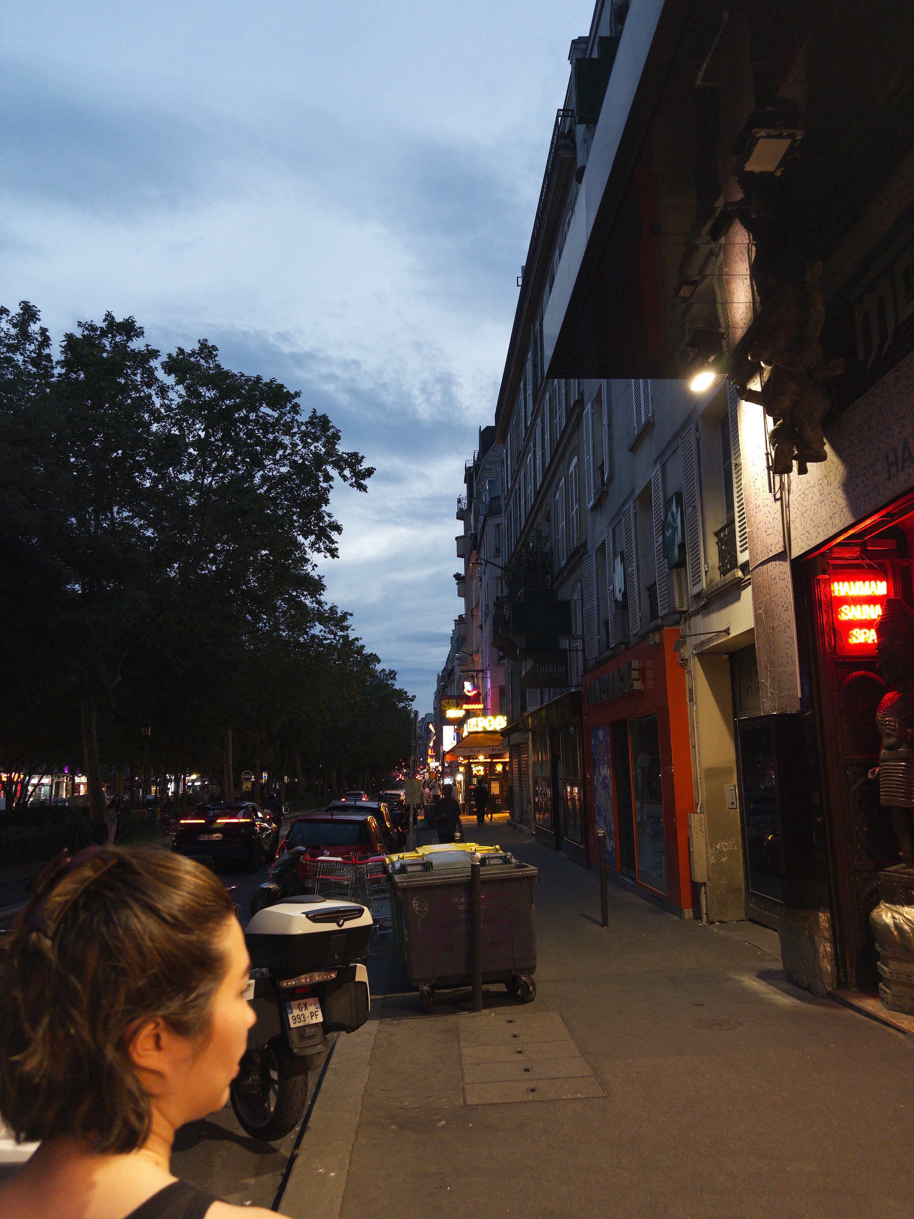 A person is walking along a dimly lit city street lined with trees, buildings, and parked cars in the evening.