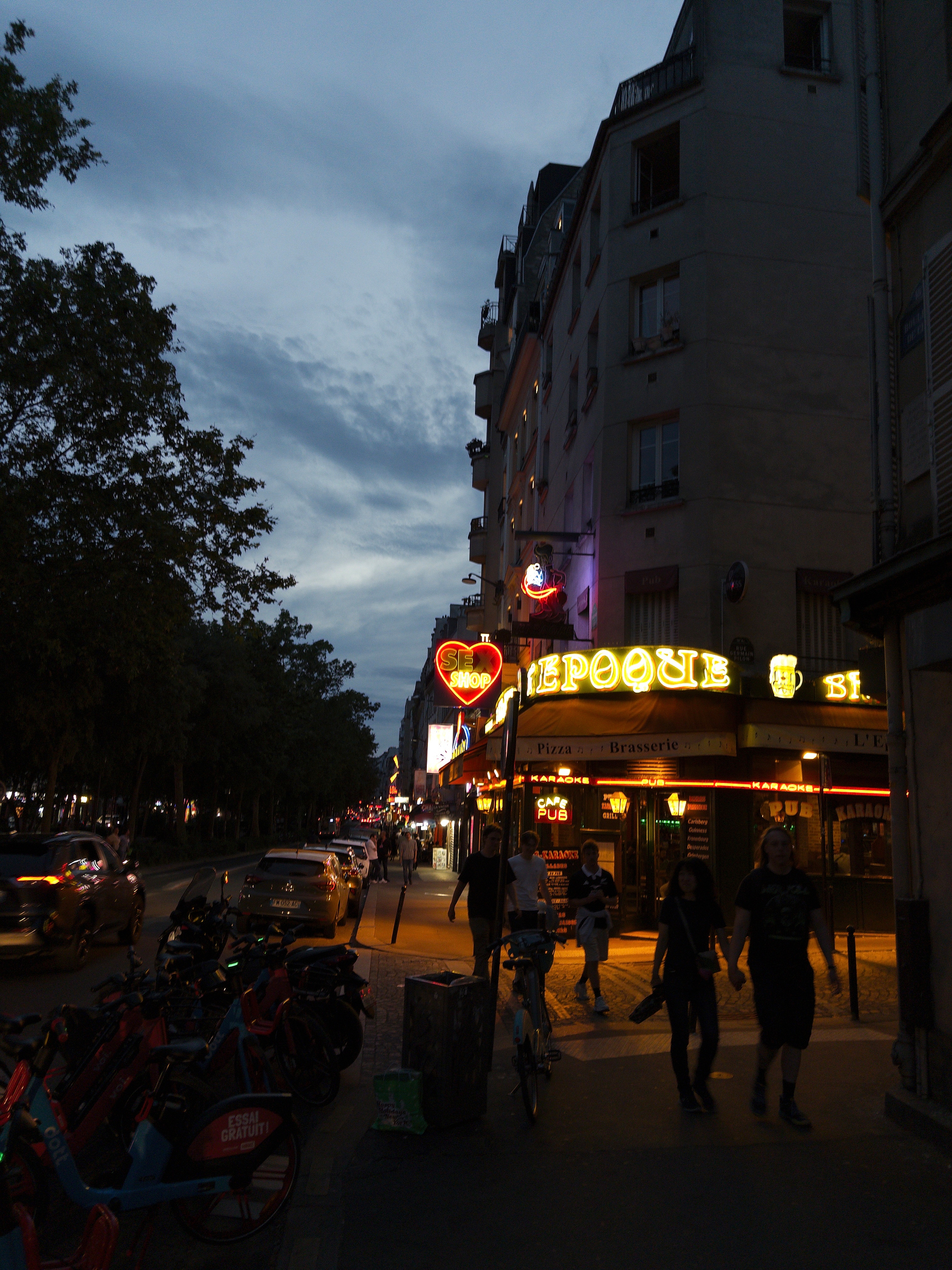 A street scene at dusk features a bustling Parisian sidewalk with neon signs illuminating a brasserie, bicycles lined up, and people walking by.