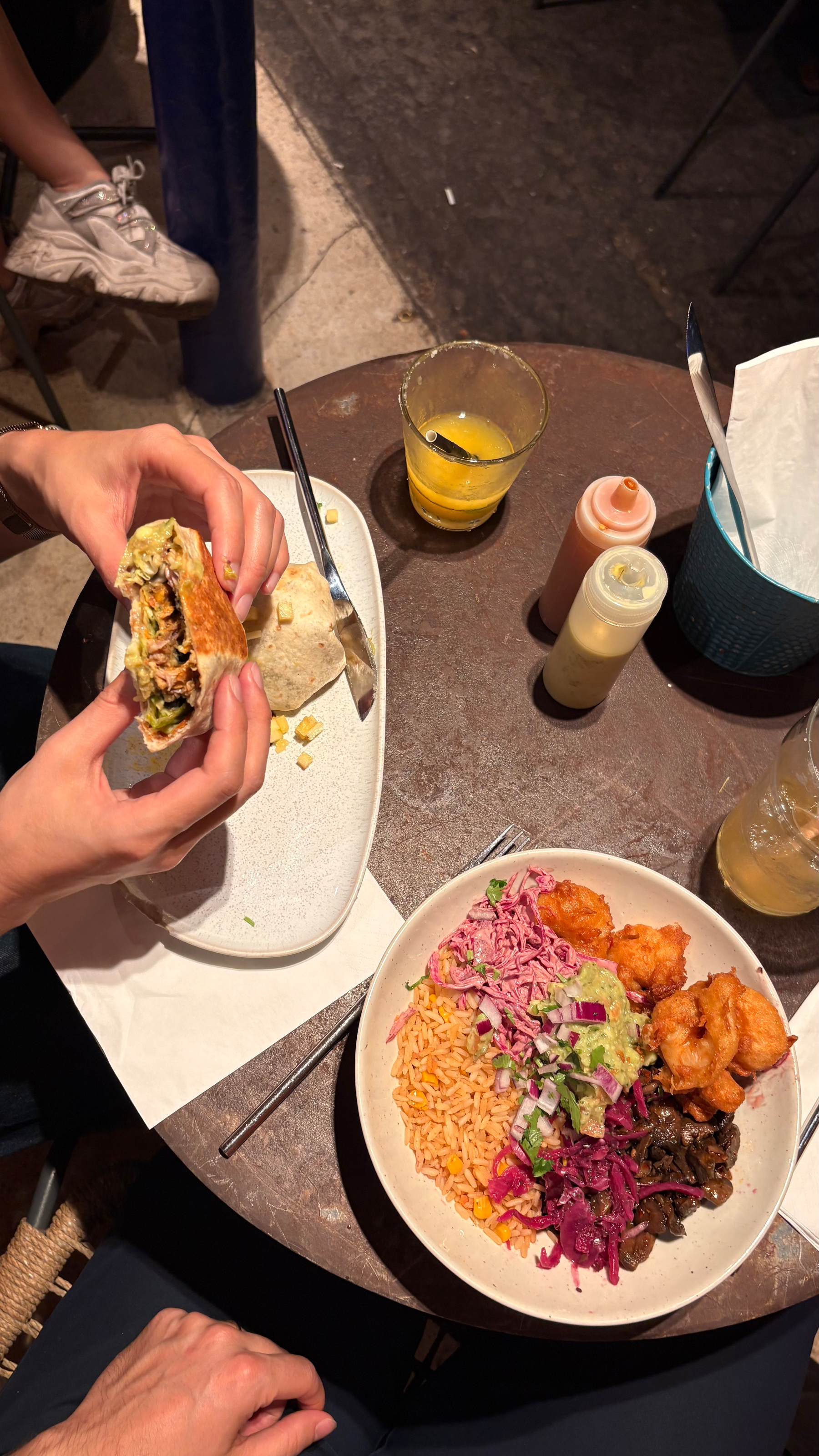 A person's hands are holding a burrito over a table with a colorful rice bowl, sauces, and a glass of frozen margarita.
