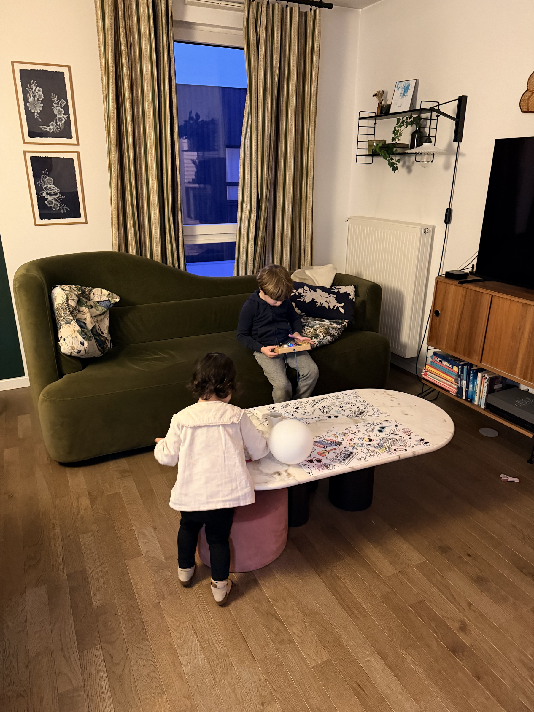A living room features a green sofa with a child sitting on it, a coffee table with artistic designs, and a toddler standing nearby holding a ball.