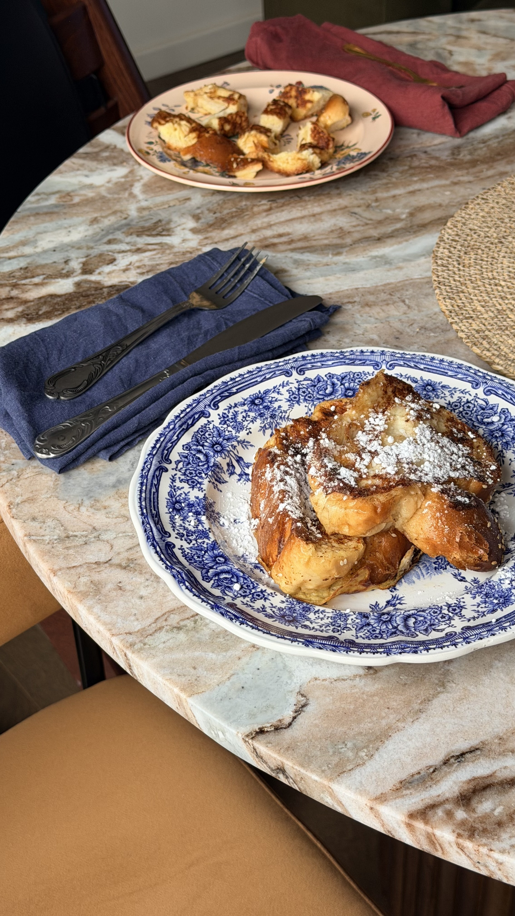 A plate of French toast dusted with powdered sugar sits on a marble table, accompanied by cutlery on a blue napkin and another dish in the background.