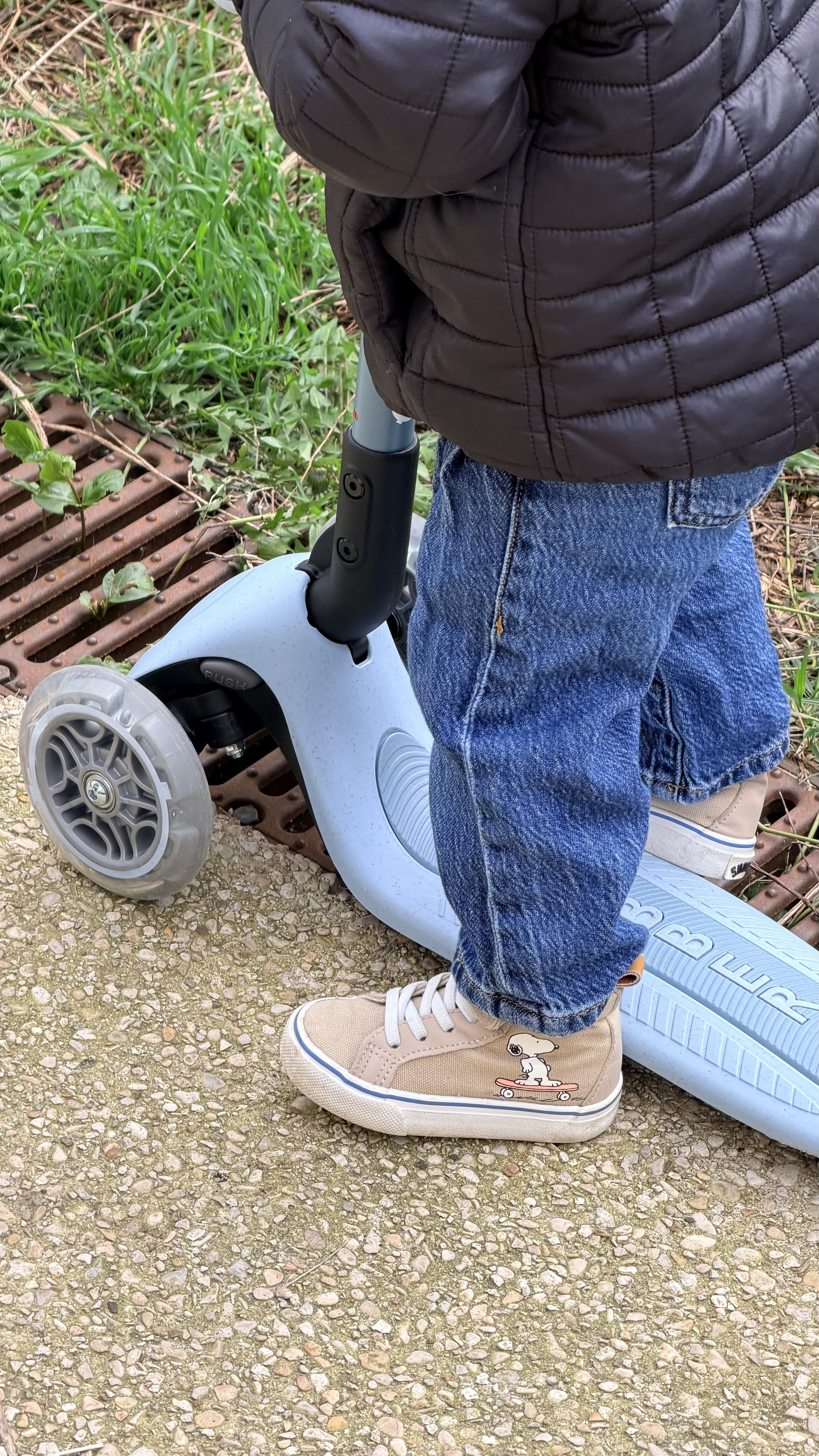 A child wearing jeans and sneakers stands next to a scooter on a pathway.