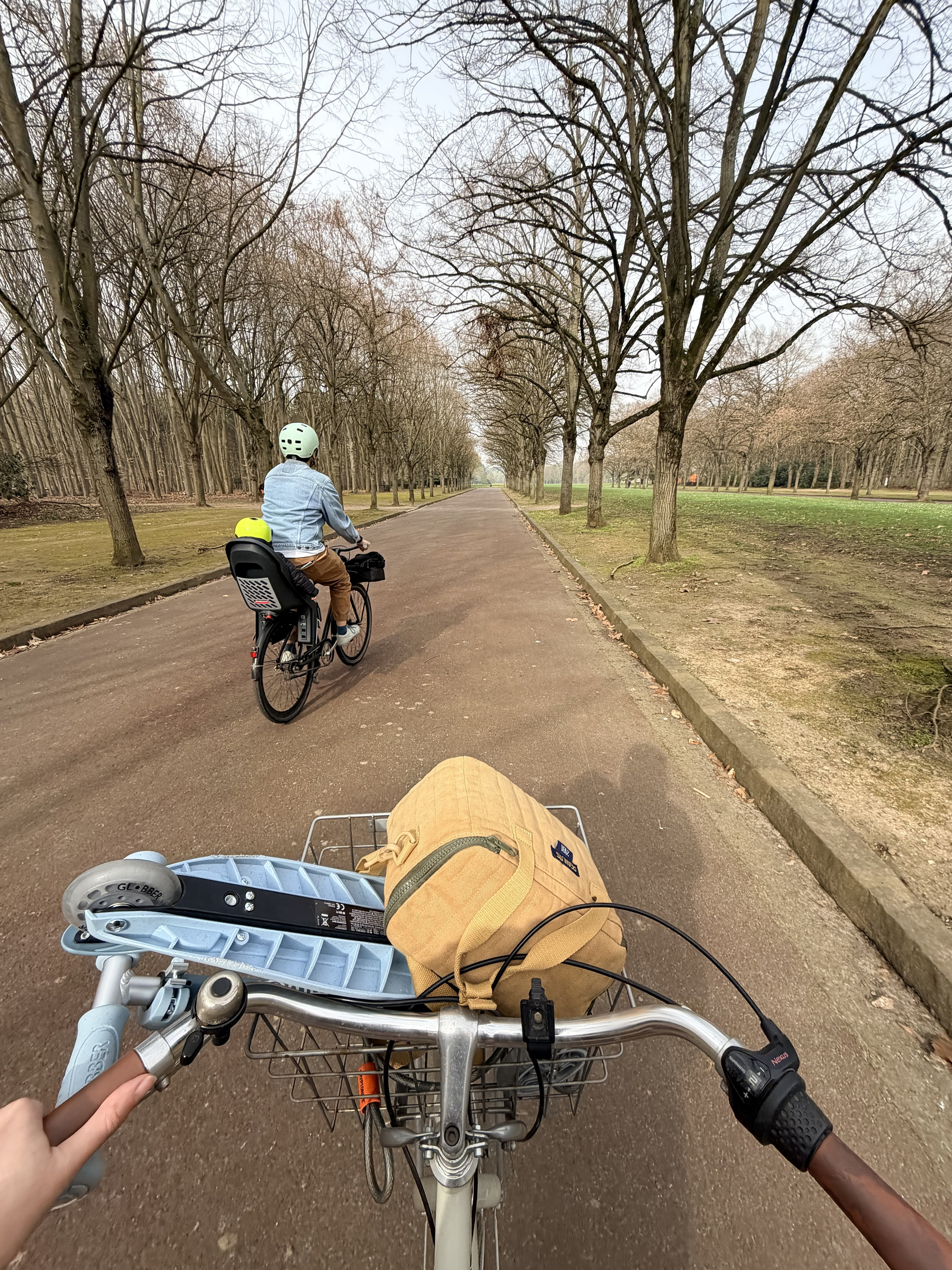 Two people are cycling down a tree-lined path, with one person in front and the other following, carrying a backpack on the bike.