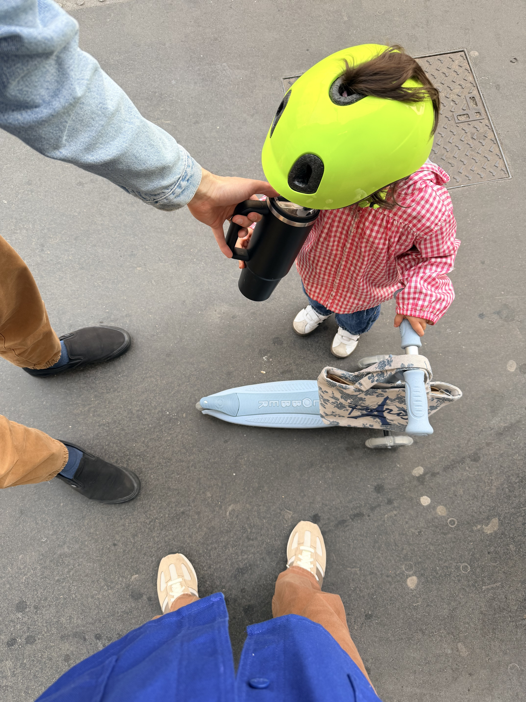 A child wearing a bright helmet and a pink checkered jacket holds a scooter while drinking from a cup, with two adults standing nearby.