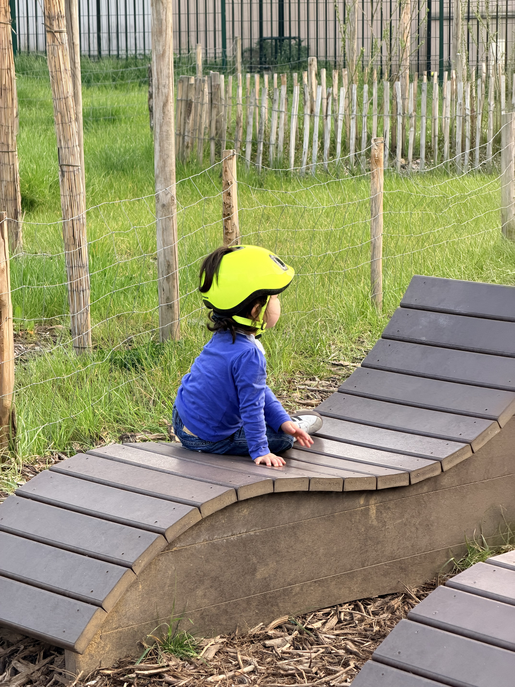 A child wearing a bright yellow helmet is sitting on a curved wooden structure in an outdoor play area.