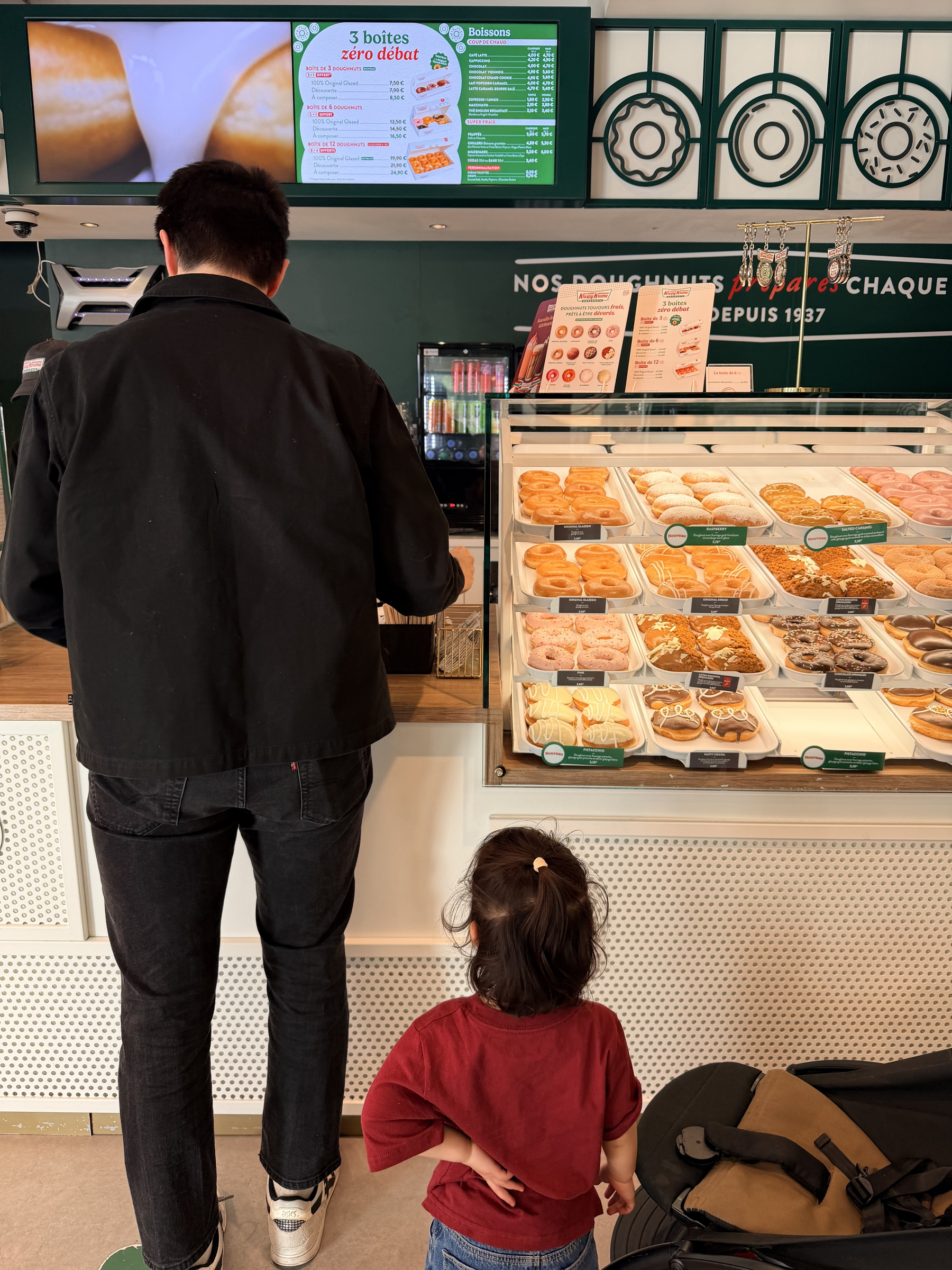 A man and a child are standing in front of a bakery display filled with various pastries and donuts.