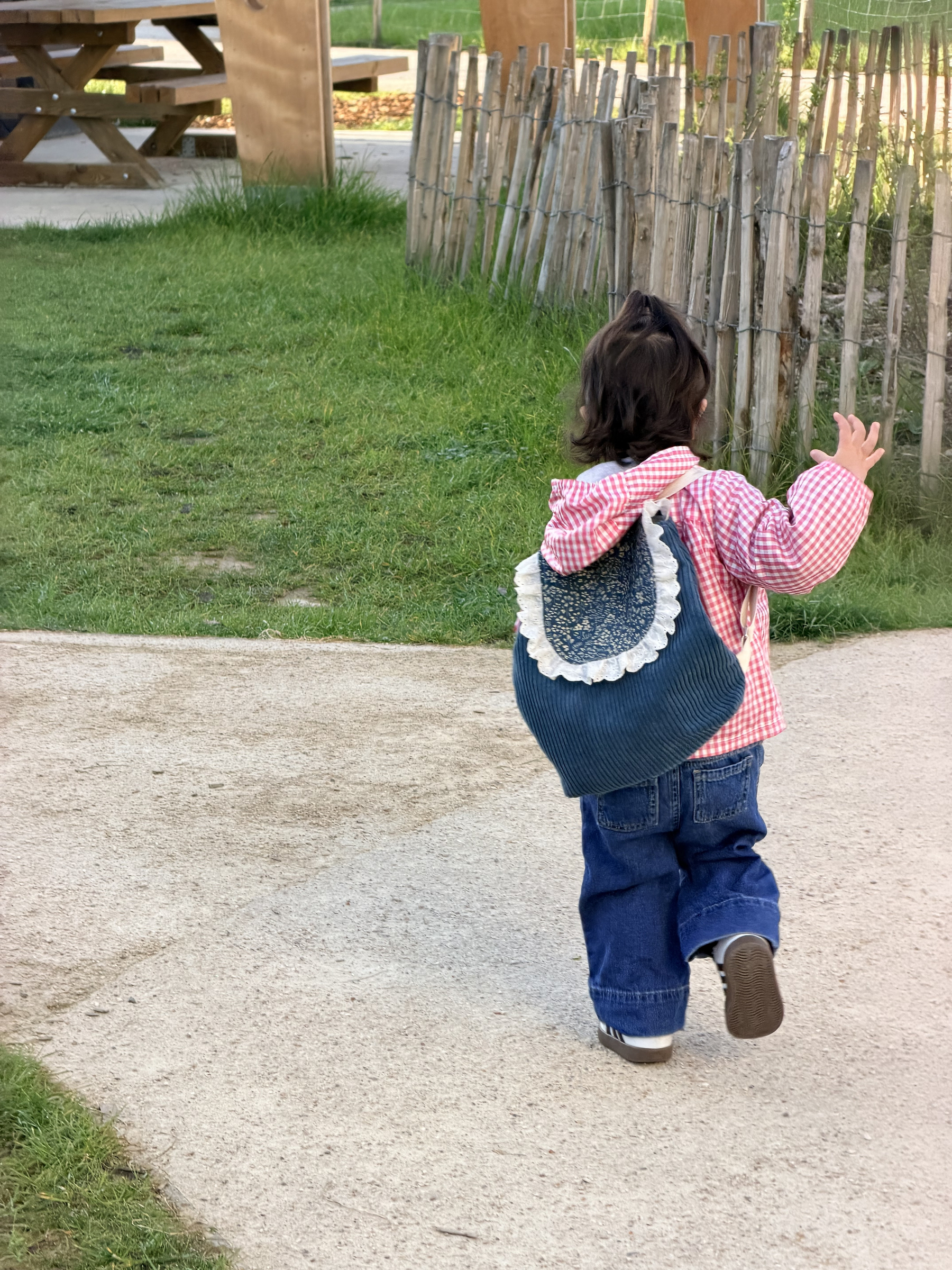 A young child wearing a red and white checkered jacket and carrying a blue backpack walks along a path near a wooden fence.