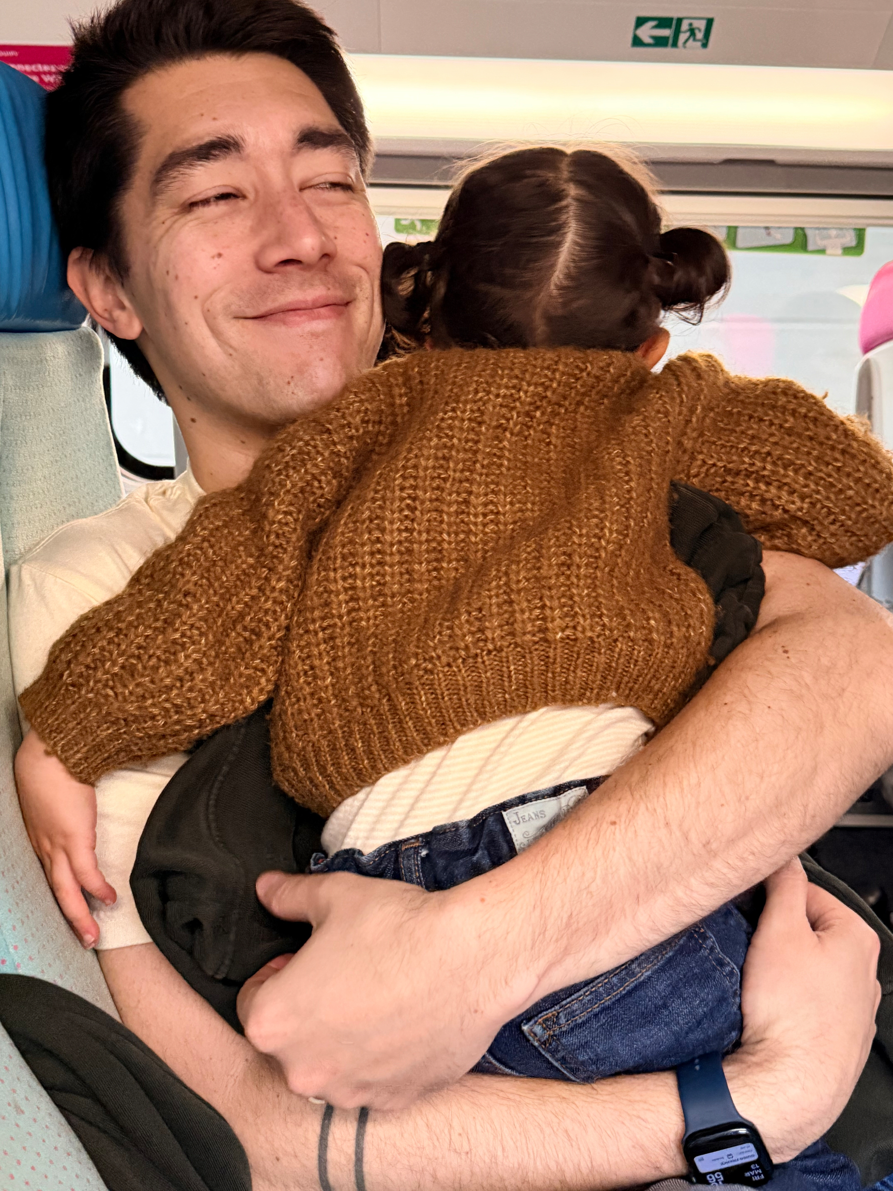 A person is joyfully holding a toddler with pigtails who is wearing a brown sweater.