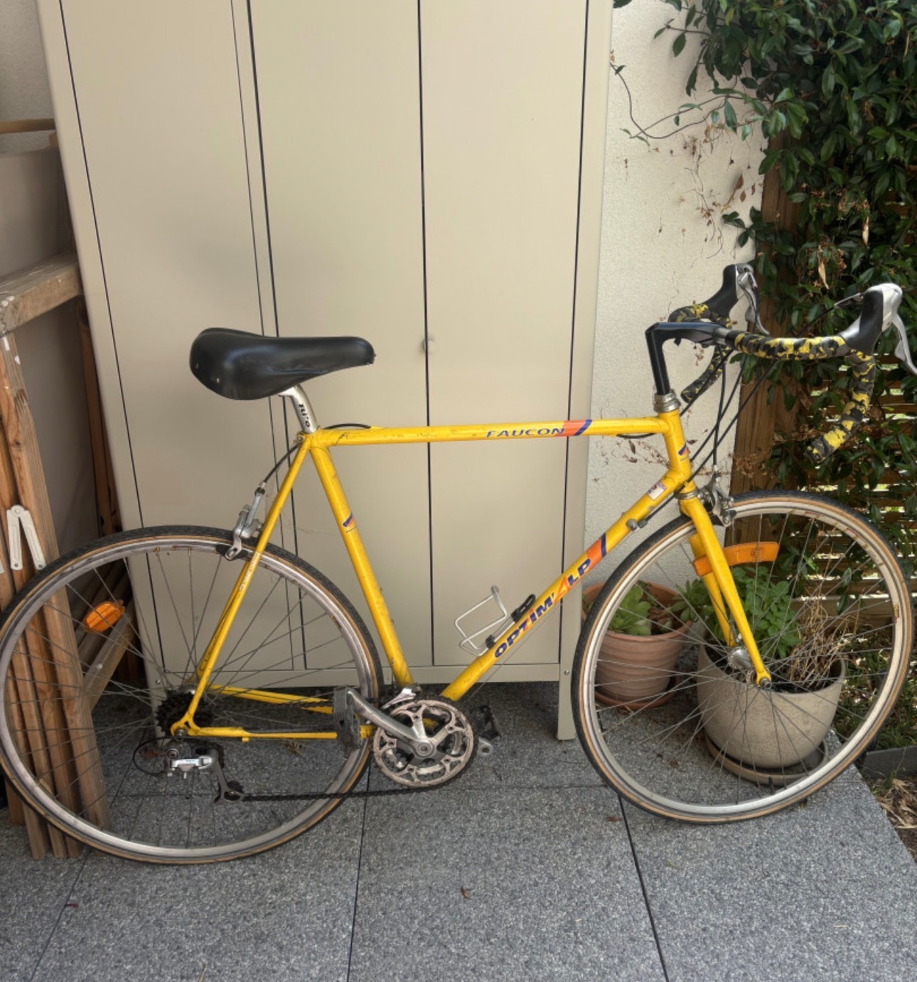 A yellow road bicycle is leaning against a light-colored cabinet outdoors next to some potted plants.