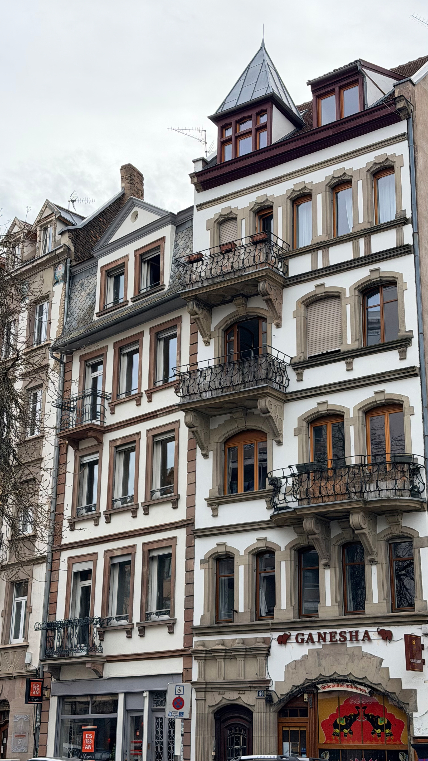 A multi-story European building features ornate balconies and window designs, with GANESHA signage visible on the ground floor.