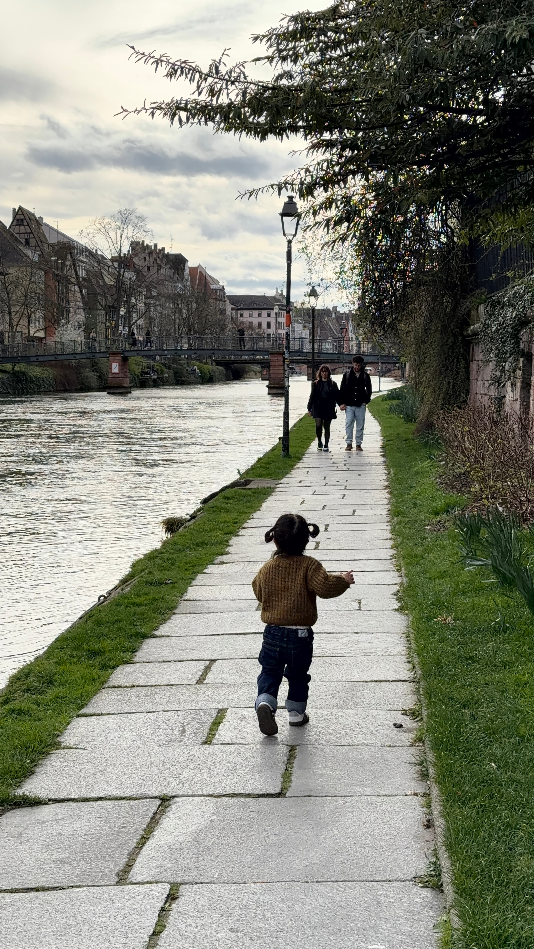A toddler walks along a riverside path while a couple strolls in the opposite direction under a cloudy sky.