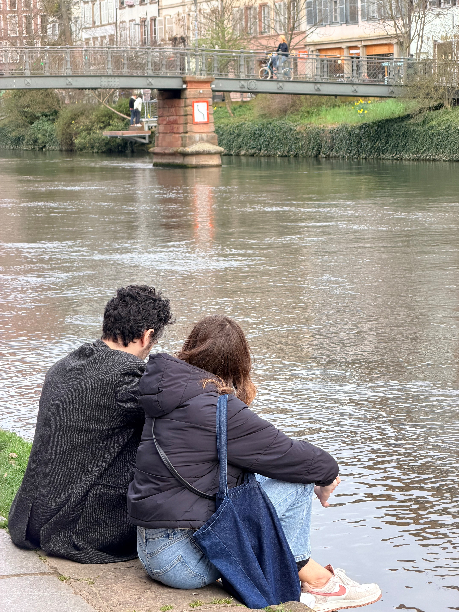 A couple sits by the riverside, overlooking a bridge with a cyclist in the background.