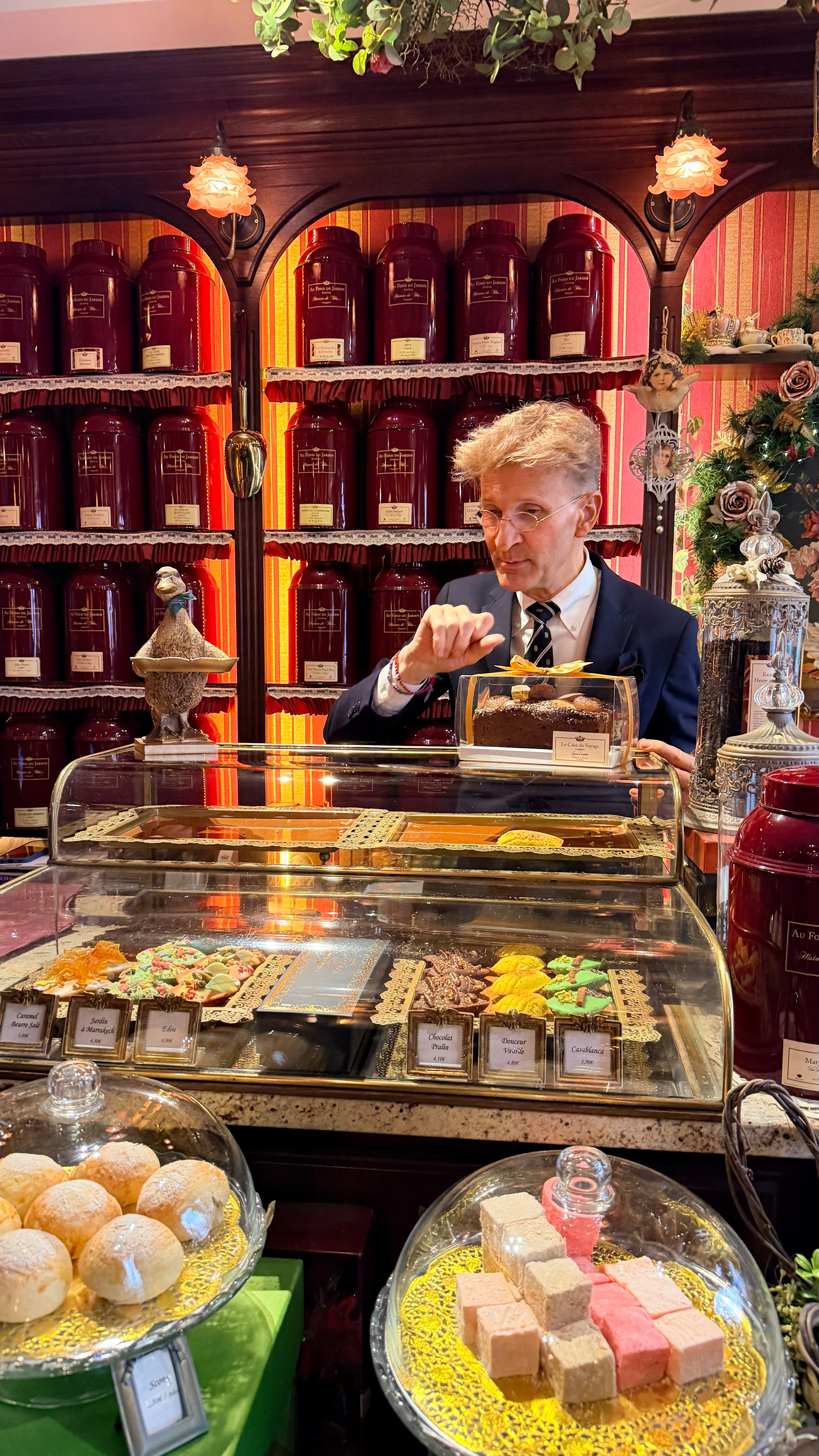 A man stands behind a counter filled with an assortment of teas and pastries in a richly decorated shop.
