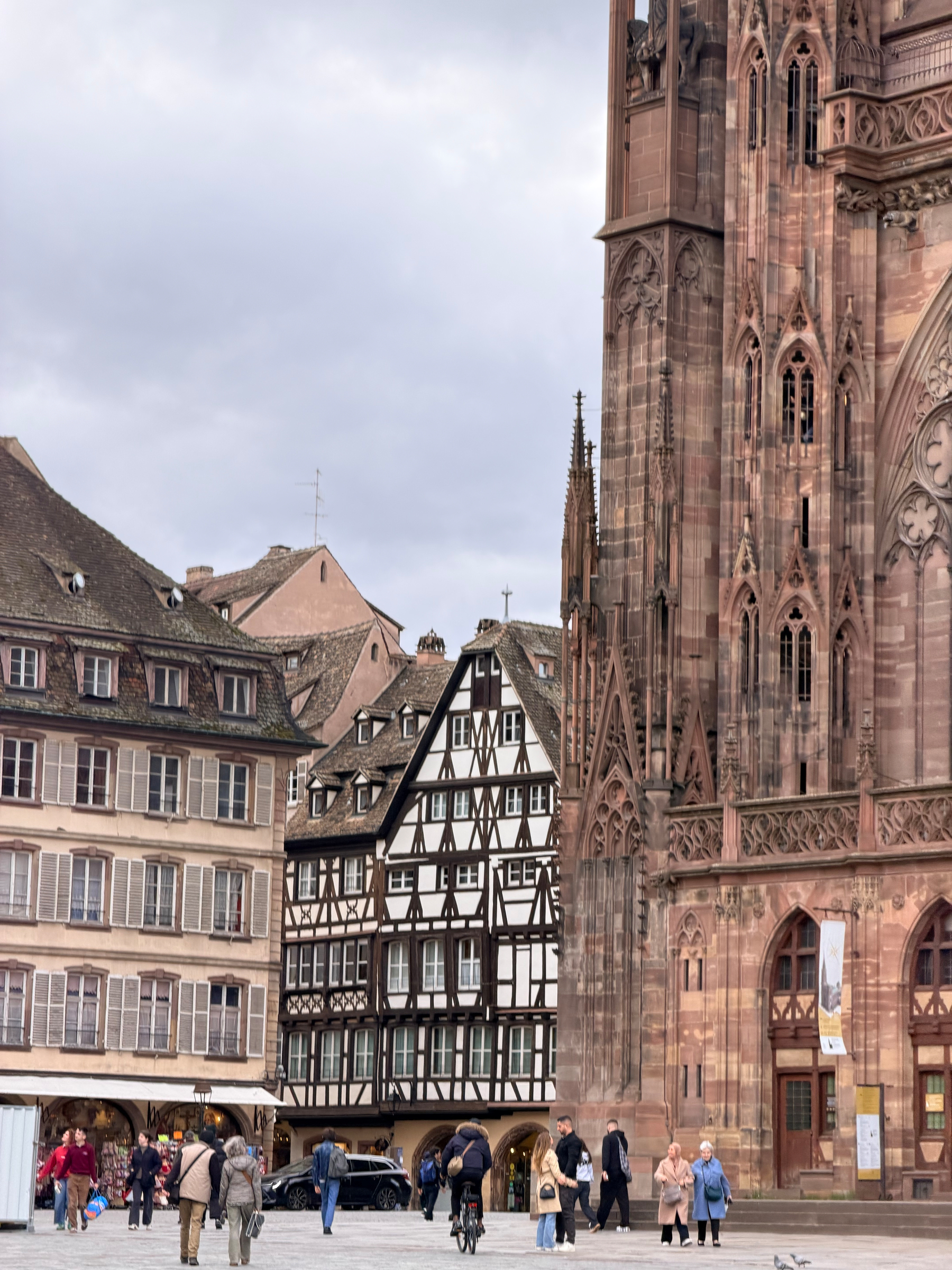 People are walking near a historic cathedral with traditional half-timbered buildings in the background.