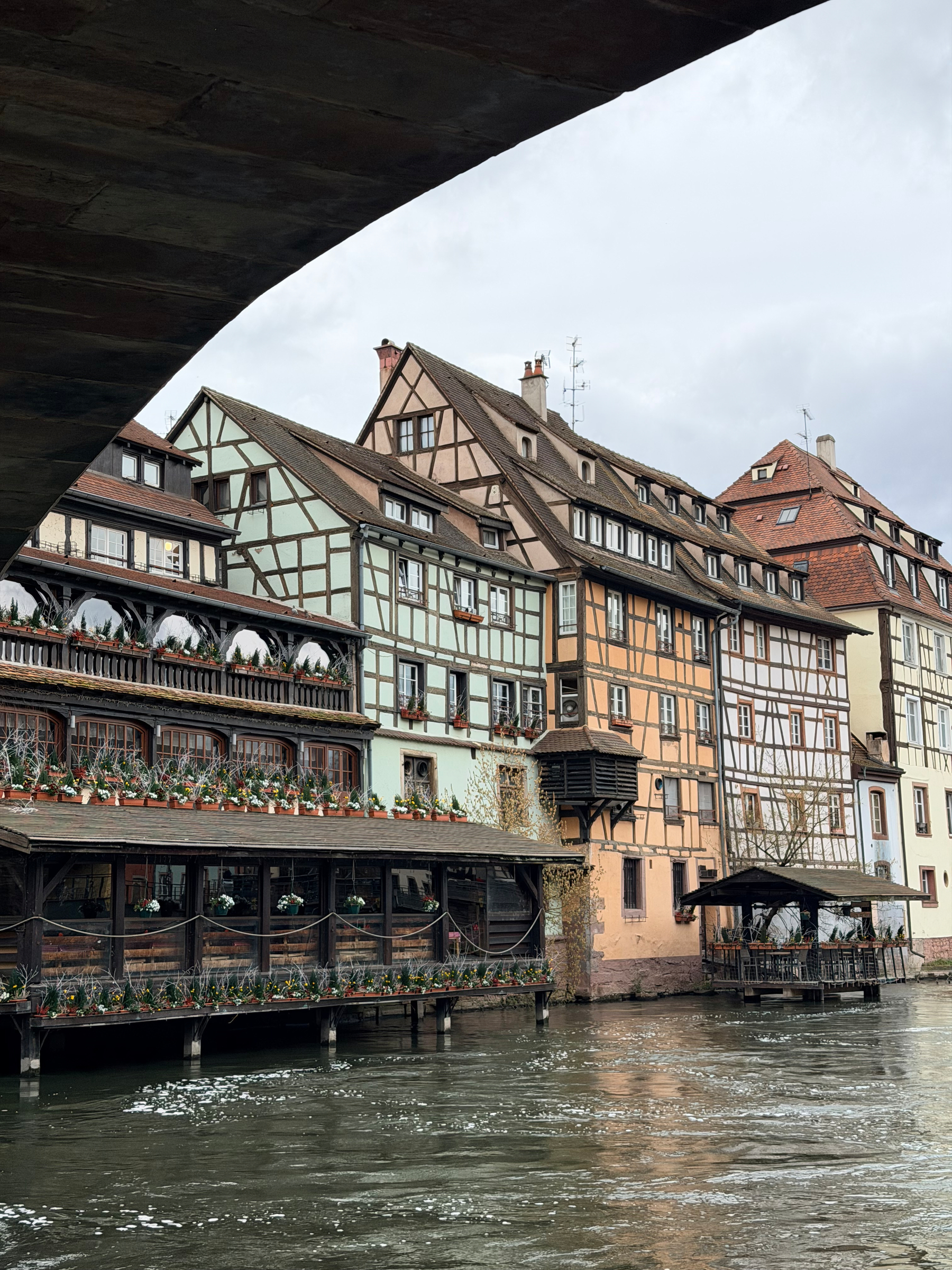 A picturesque riverside scene features half-timbered buildings adorned with flowers under a cloudy sky.
