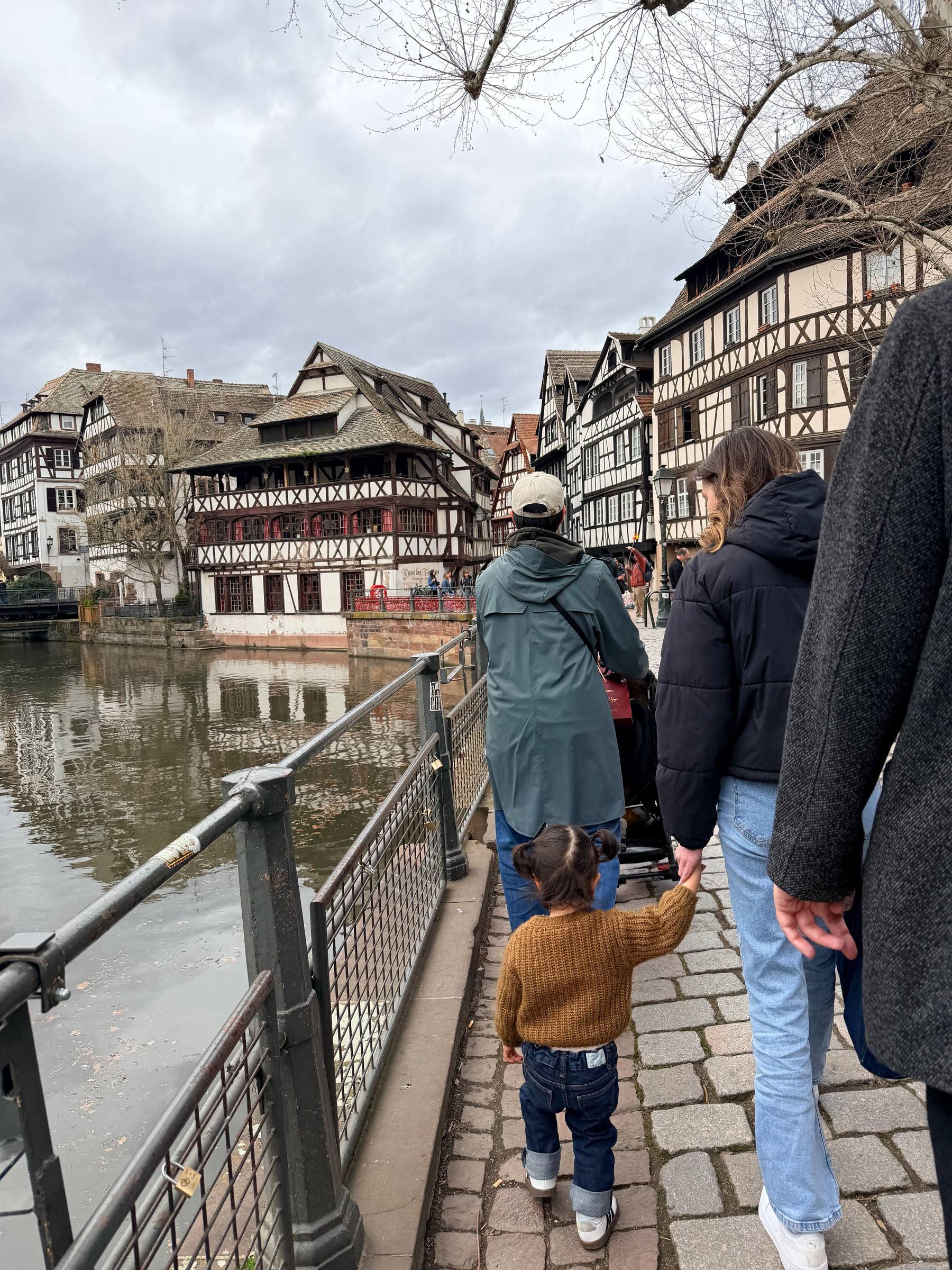 A group of people walk along a riverside path lined with traditional half-timbered houses in a picturesque setting.