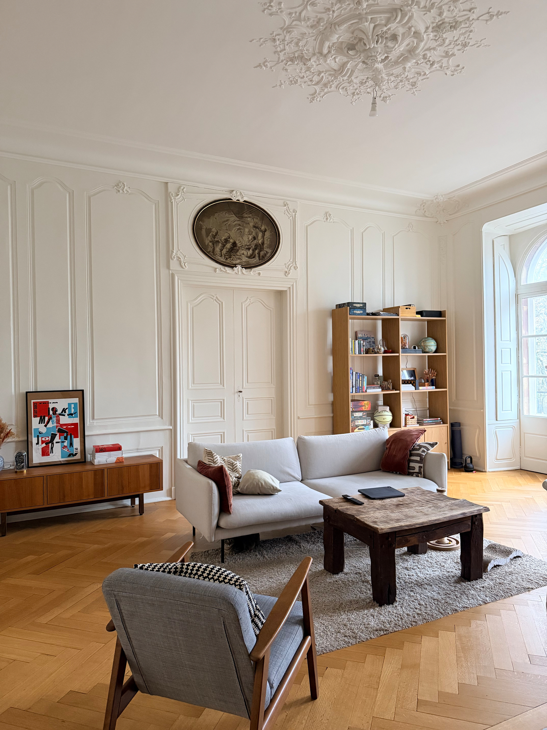 A stylish living room features a white sofa, a wooden coffee table on a rug, a bookshelf, and an ornate ceiling.