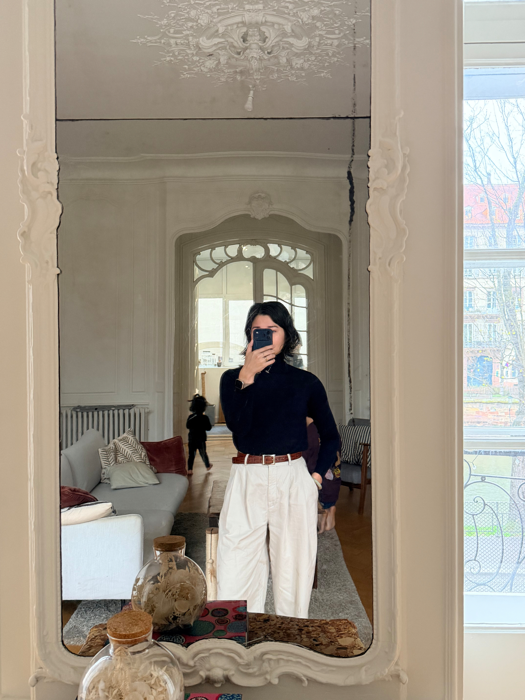 A person is taking a mirror selfie in a stylish, elegantly decorated room featuring ornate moldings and a glimpse of a cozy living space.