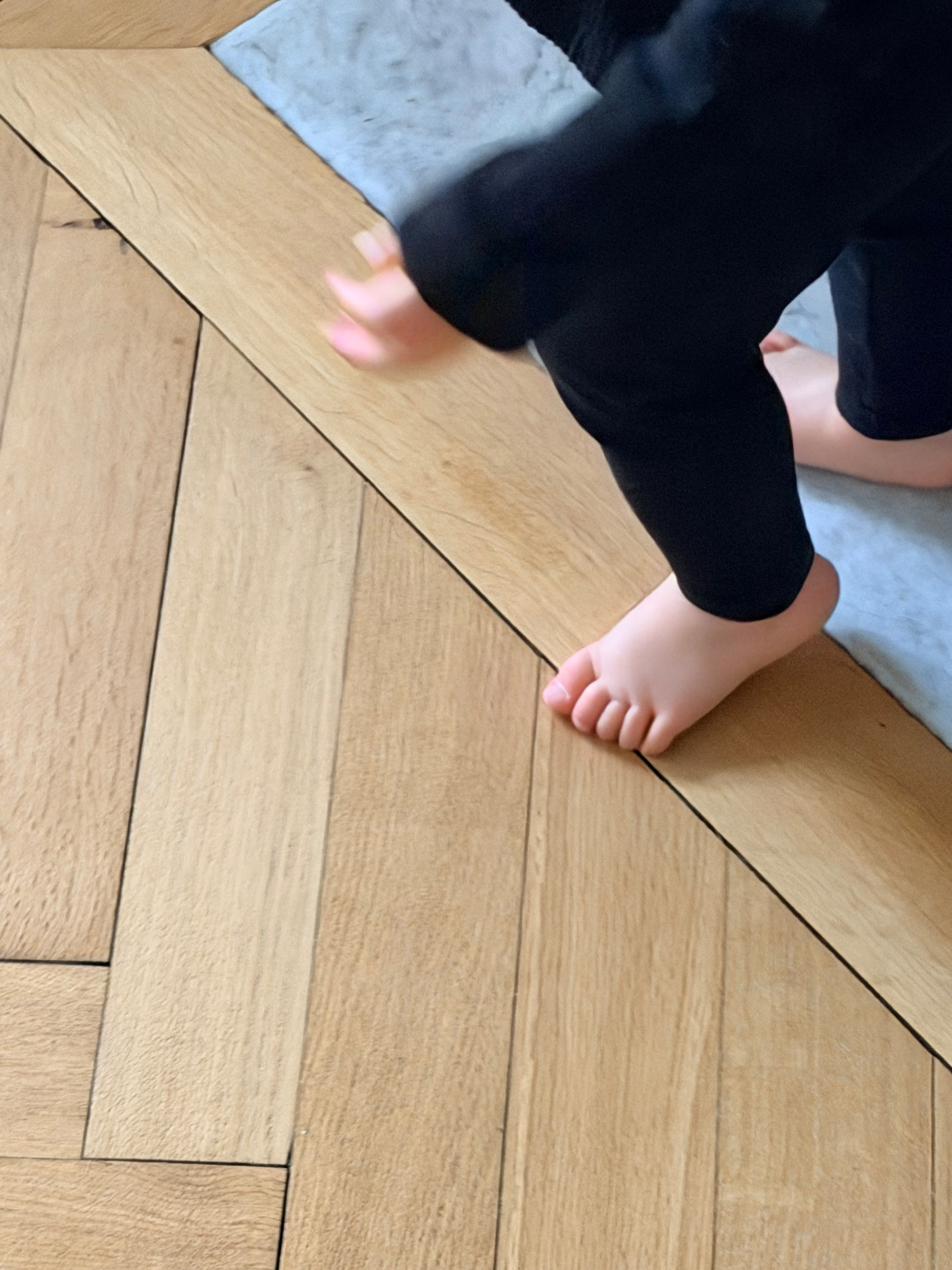 A child wearing black pants is walking on a wooden herringbone floor.
