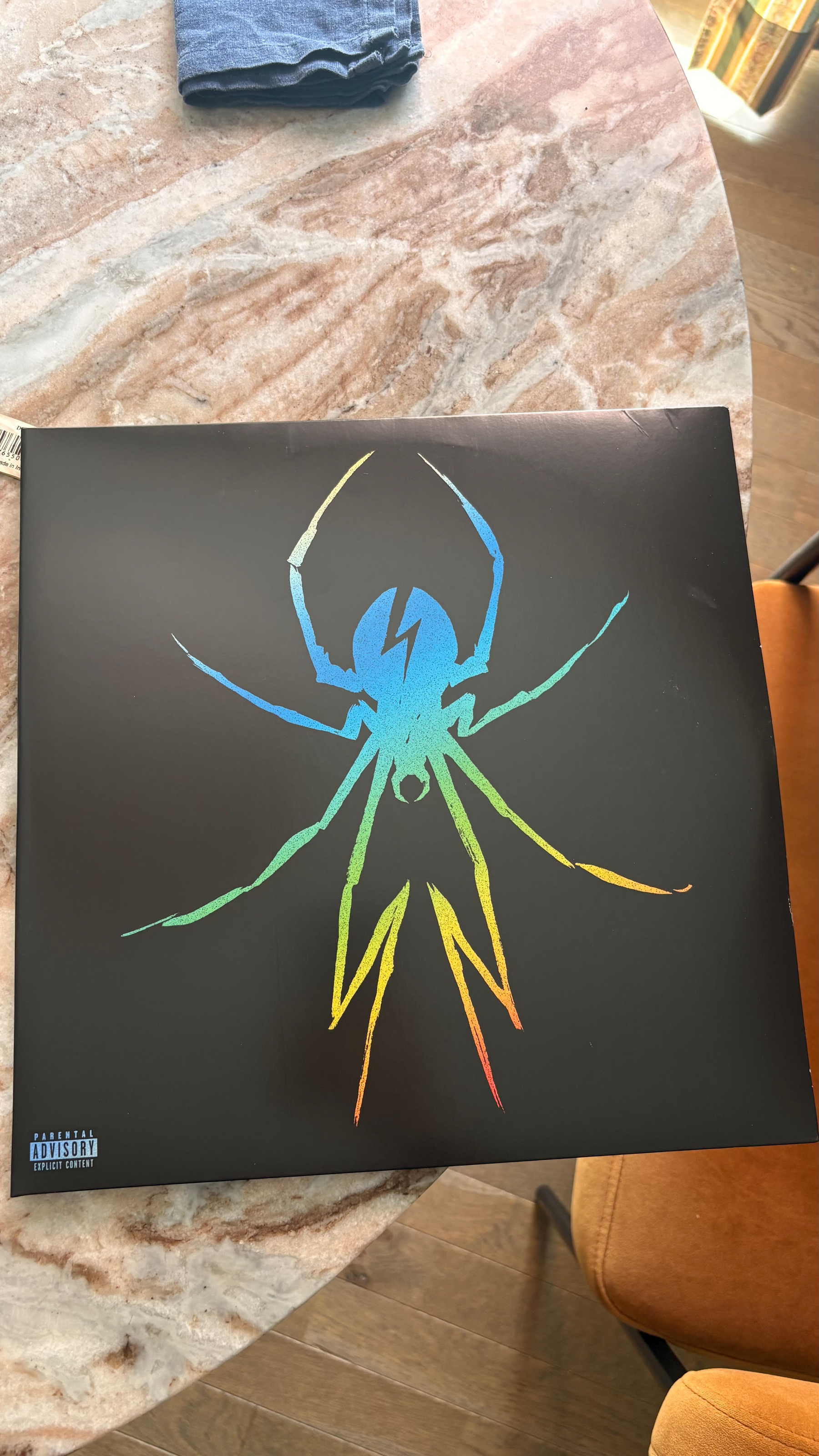 A black square cover features a colorful spider with a lightning bolt on its back and a parental advisory warning in the bottom left corner.