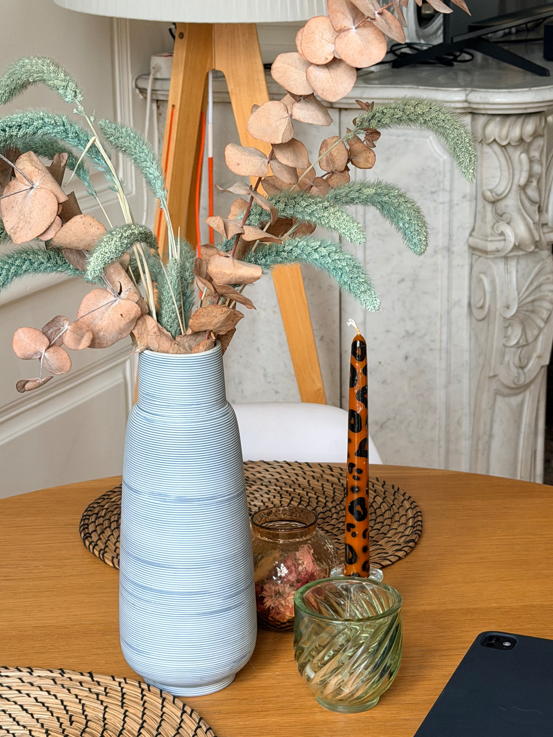 A vase with dry flowers and grasses sits on a wooden table next to decorative glassware and a candle, with a fireplace in the background.